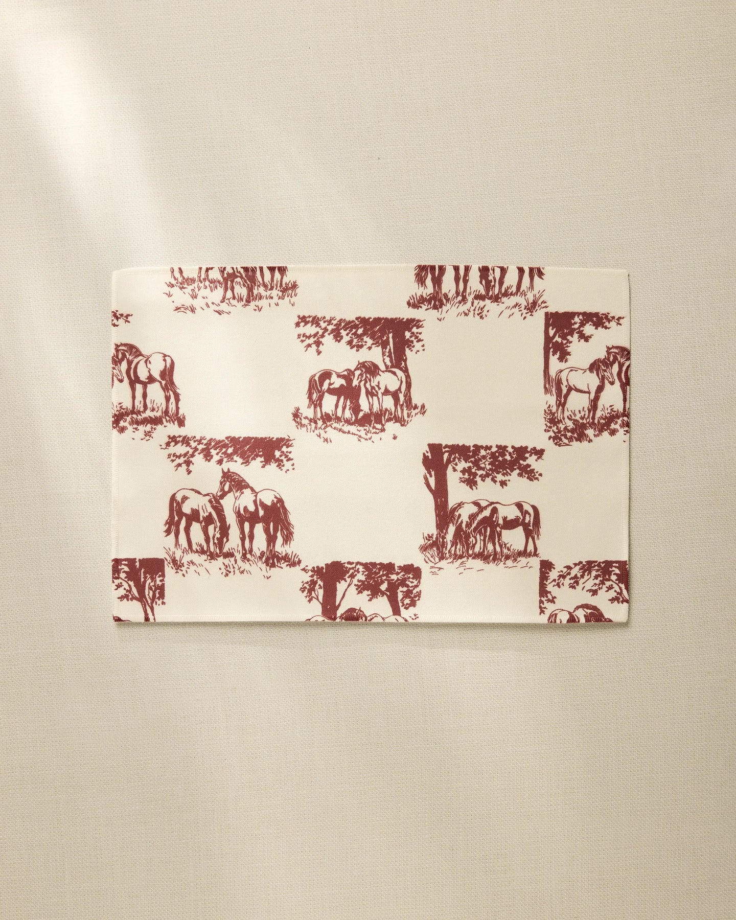 The Geometry Horse Check Cream is a rectangular, cream-colored, machine washable placemat featuring maroon horses and trees in a repeating pattern, shown on a neutral, lightly textured surface.