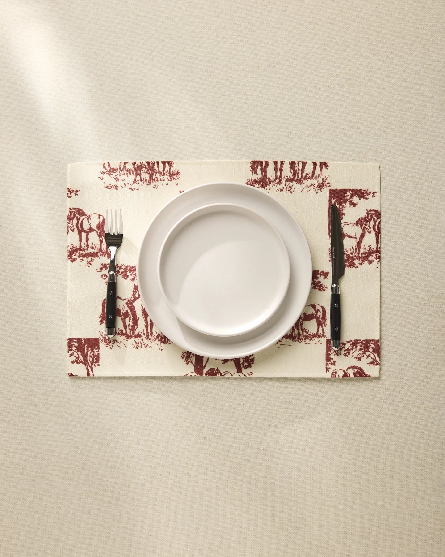 A white plate sits on the Geometry Horse Check Cream placemat with maroon horse designs, flanked by a fork and knife on a beige tablecloth. These machine-washable placemats blend style and easy care for any setting.