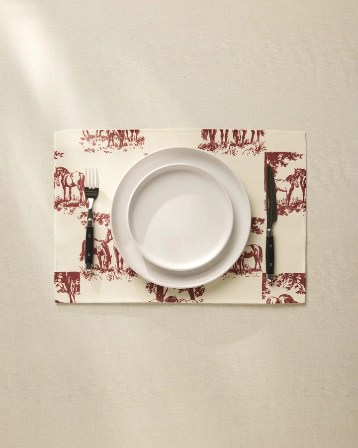 A white plate sits on the Geometry Horse Check Cream placemat with maroon horse designs, flanked by a fork and knife on a beige tablecloth. These machine-washable placemats blend style and easy care for any setting.