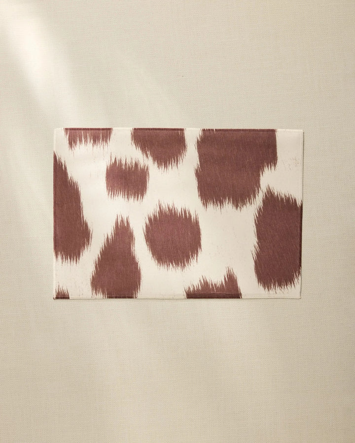 A rectangular Rodeo envelope by Geometry, featuring a brown and white animal print, rests on light beige fabric as sunlight casts a soft shadow—reminiscent of the brand’s stylish placemats.