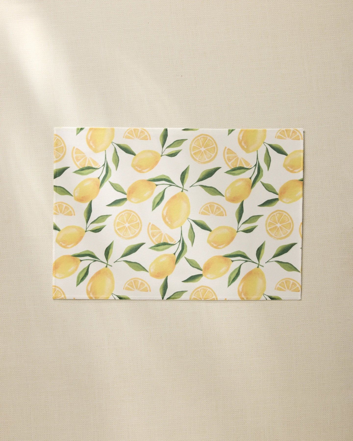 The Geometry Limoncello rectangular white recycled placemat features a pattern of whole lemons, lemon slices, and green leaves on a light beige textured surface.