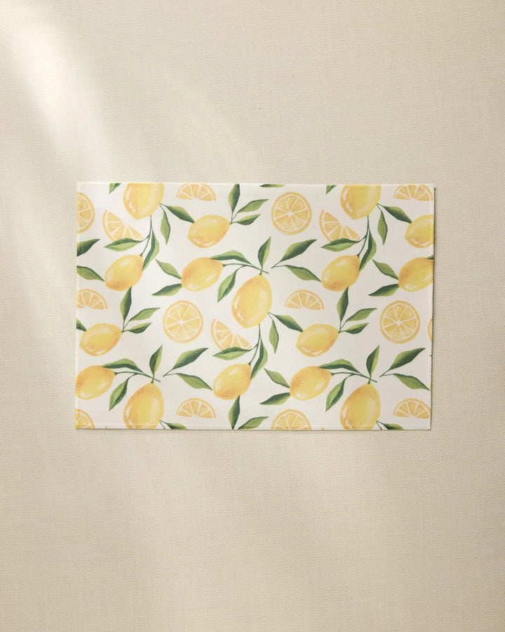 The Geometry Limoncello rectangular white recycled placemat features a pattern of whole lemons, lemon slices, and green leaves on a light beige textured surface.