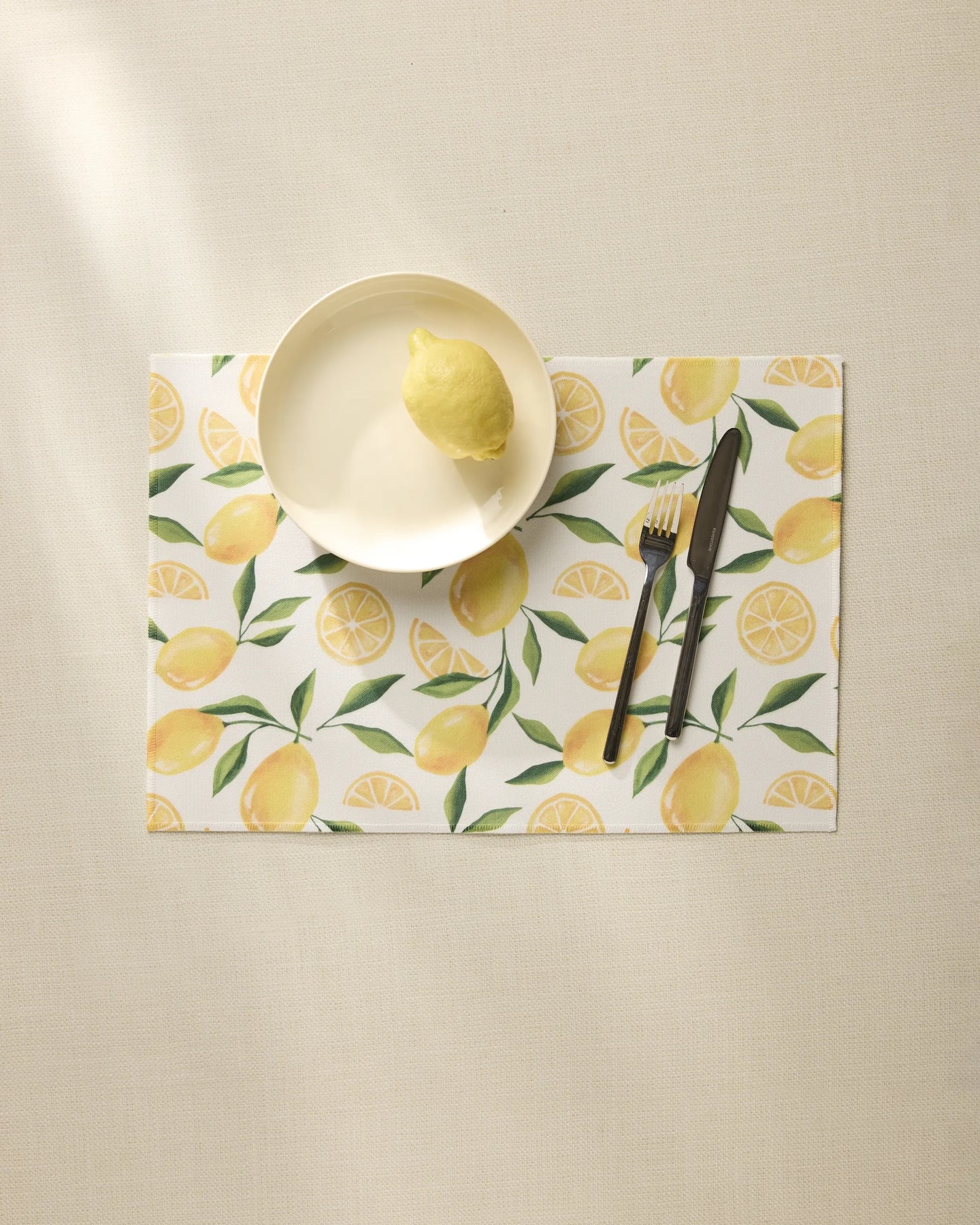 A cream plate with a lemon rests on the Geometry Limoncello placemat, featuring a vibrant lemon pattern. A knife and fork are placed to the right, all set atop a light beige tablecloth.