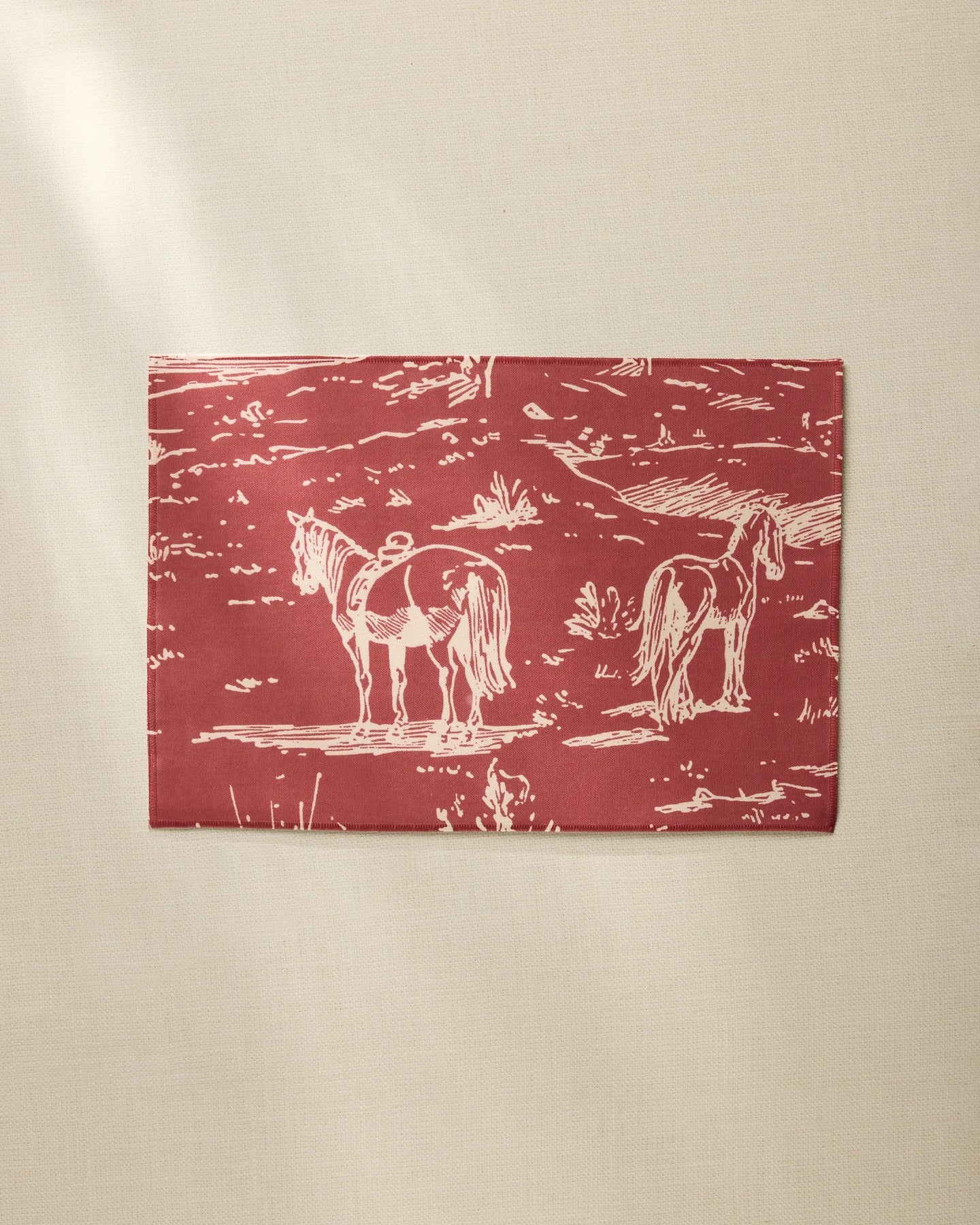The Geometry Western Scene placemat is rectangular, red, and made from recycled materials, featuring a white line drawing of two horses in a field, shown on a beige surface.