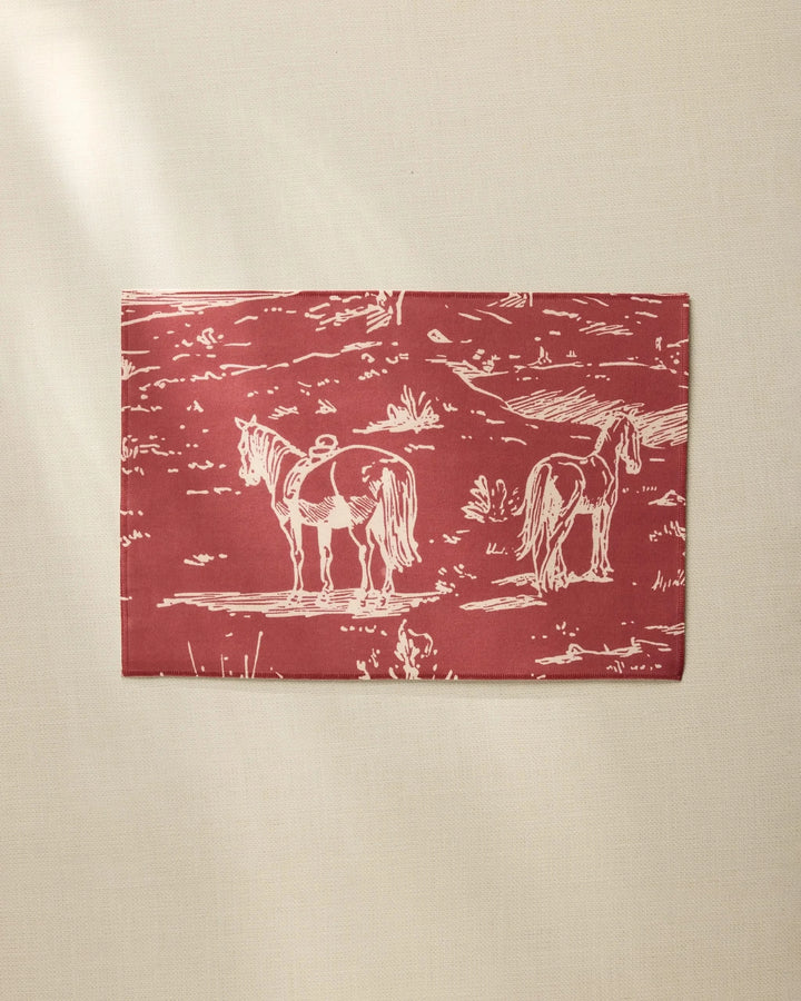 The Geometry Western Scene placemat is rectangular, red, and made from recycled materials, featuring a white line drawing of two horses in a field, shown on a beige surface.