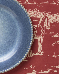 A blue ceramic plate with a beaded edge rests on the Geometry Western Scene placemat—a red, machine-washable tablecloth adorned with a white line drawing of a horse and landscape.