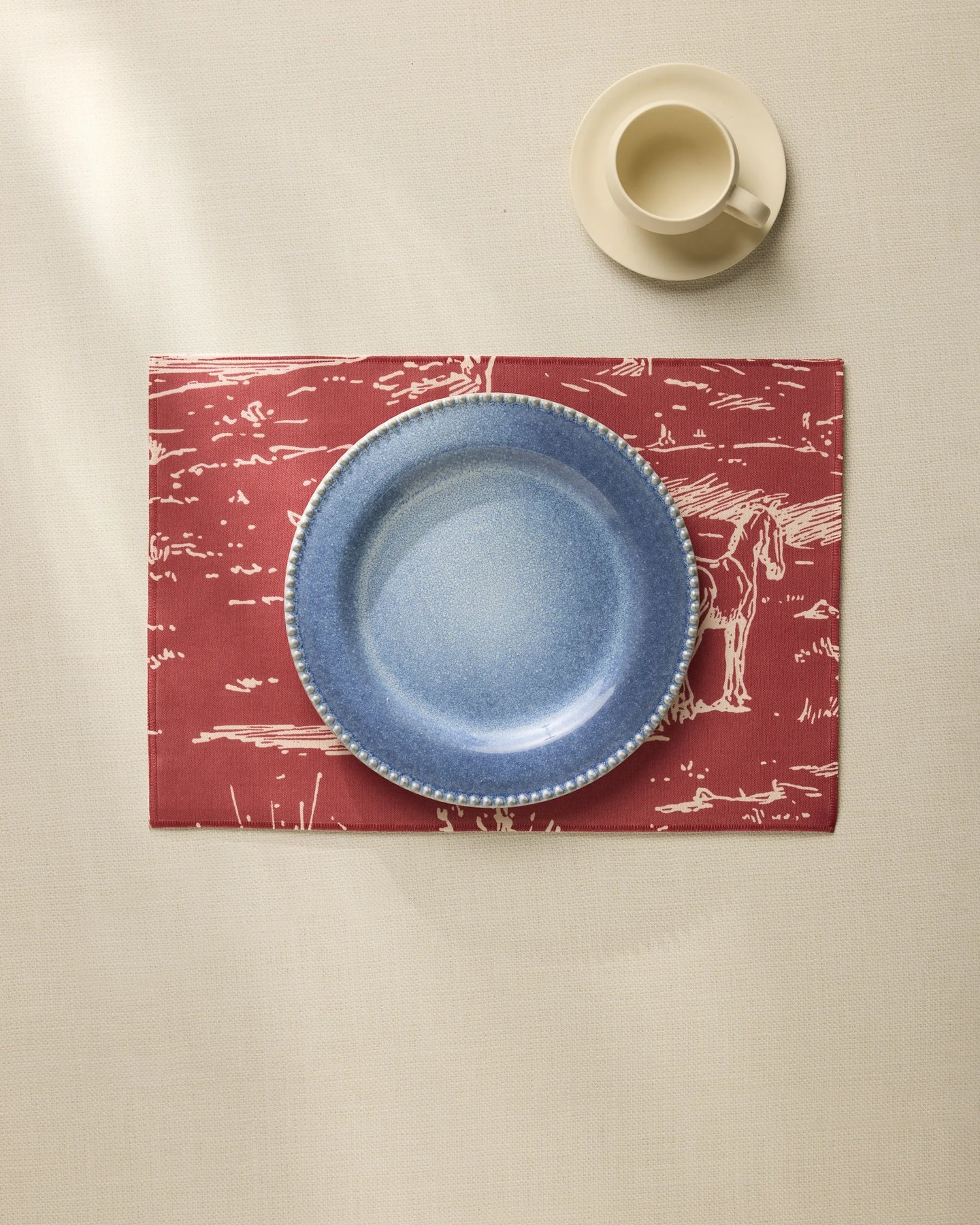 The Geometry Western Scene set features a blue ceramic plate on a machine washable red placemat with white horse sketches, paired with a cream-colored cup and saucer, all displayed atop a light tablecloth.