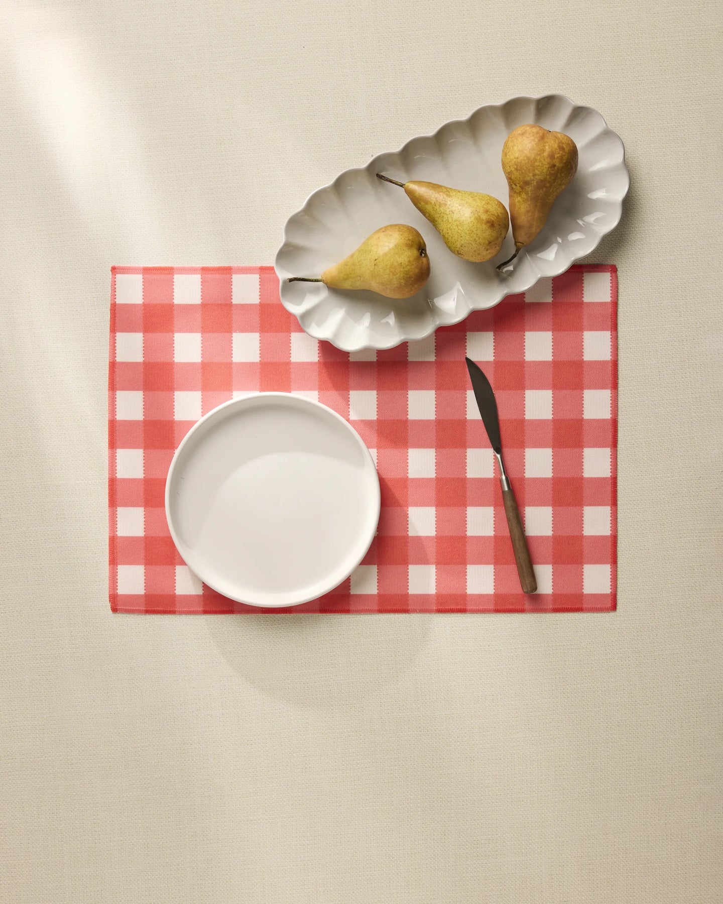 A white bowl, a knife, and a scalloped dish with three pears are arranged on Geometry’s Ripe Tomato Gingham placemat atop a light-colored tablecloth.