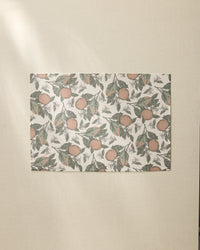 The Valencia rectangular sheet by Geometry, featuring an orange and green leaf pattern like recycled placemats, rests on a beige textured surface bathed in soft natural light from the left.