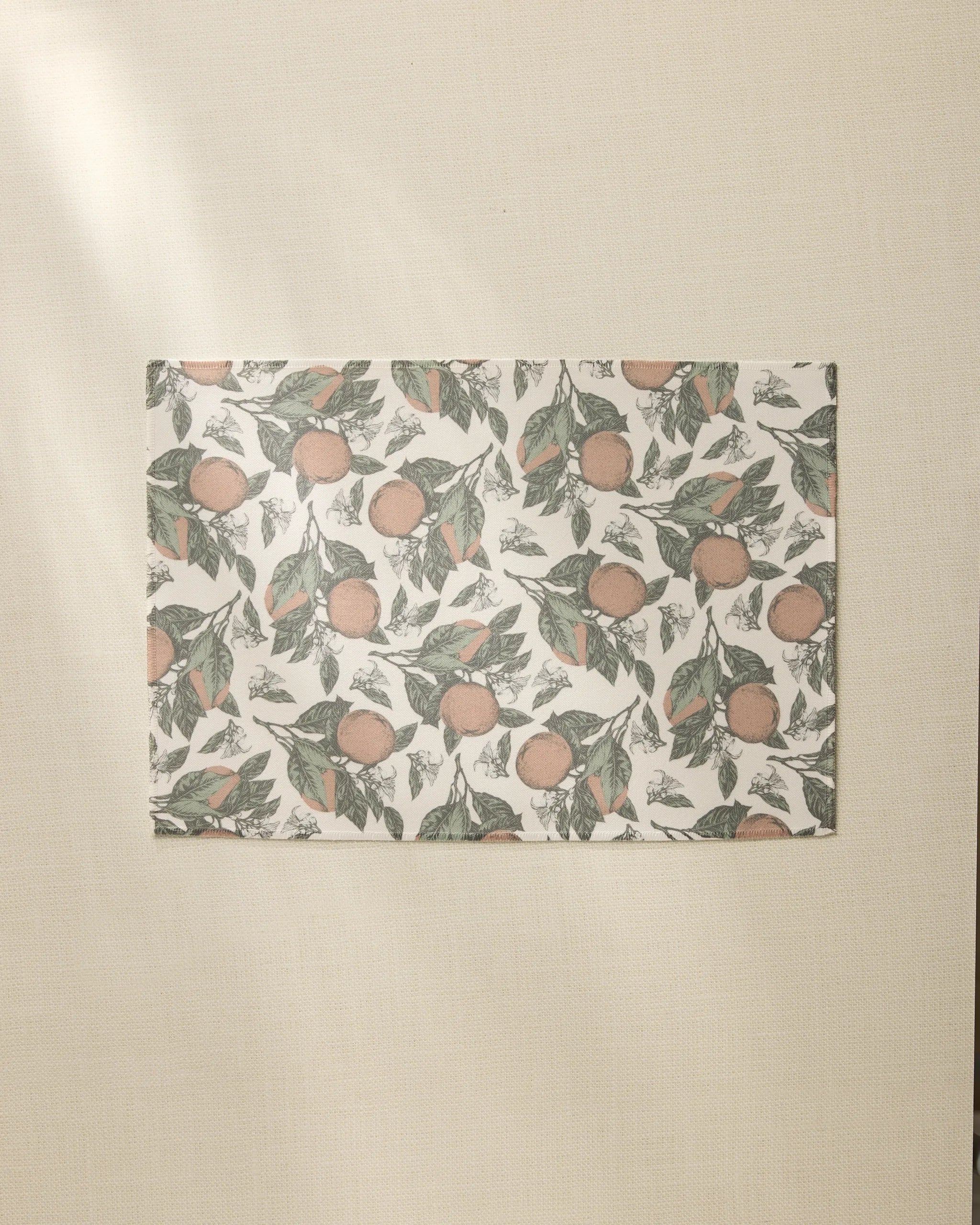 The Valencia rectangular sheet by Geometry, featuring an orange and green leaf pattern like recycled placemats, rests on a beige textured surface bathed in soft natural light from the left.