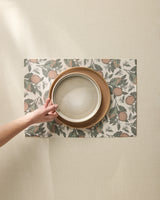 A hand sets a light gray Valencia bowl atop two brown plates on Geometrys machine washable placemats with an orange fruit and leaf pattern, arranged on a beige textured surface.