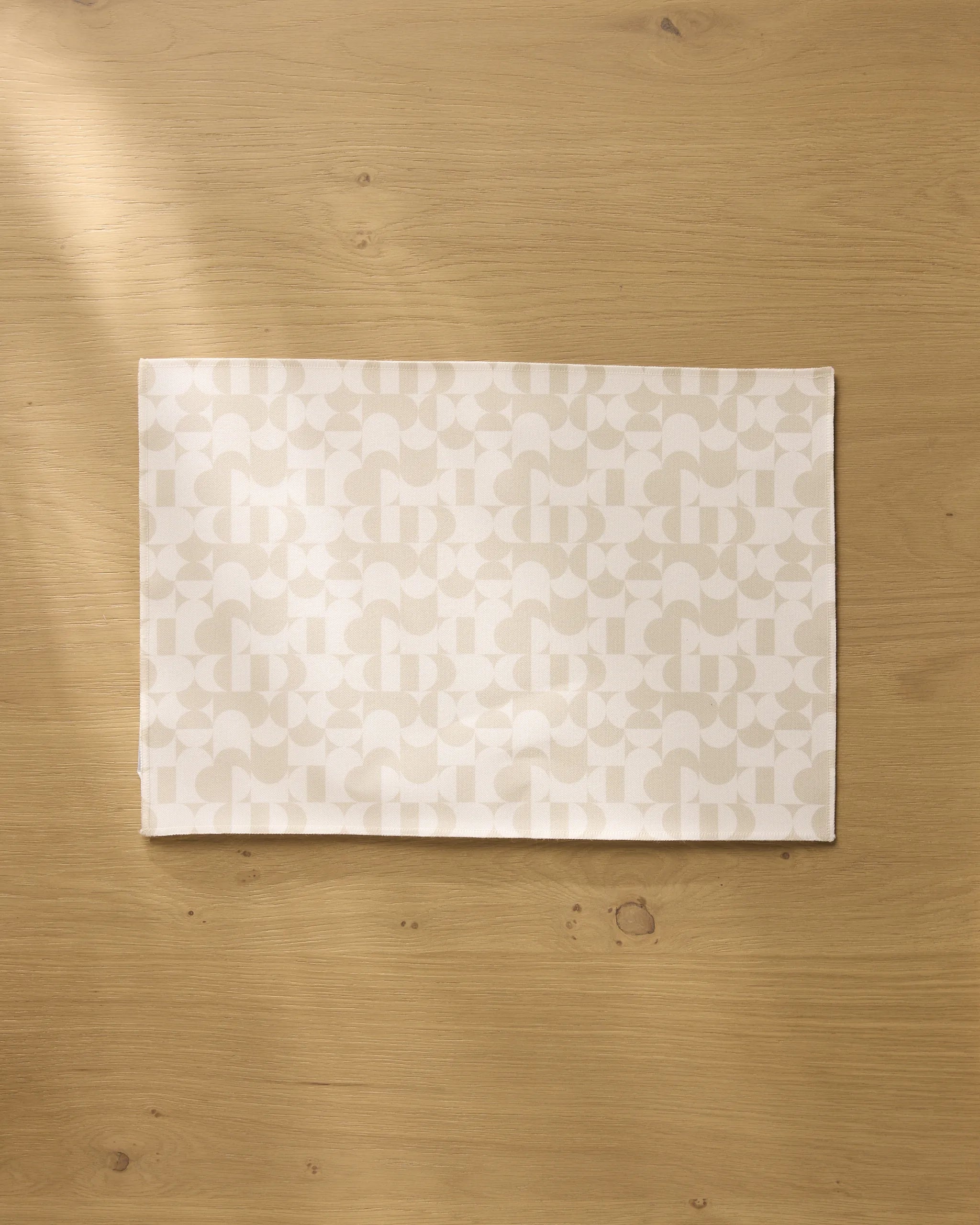 The Geometry Circle Playground placemat, with its subtle geometric design, is centered on a light wood table. Soft light casts gentle shadows, highlighting the elegant and machine washable style of these placemats.