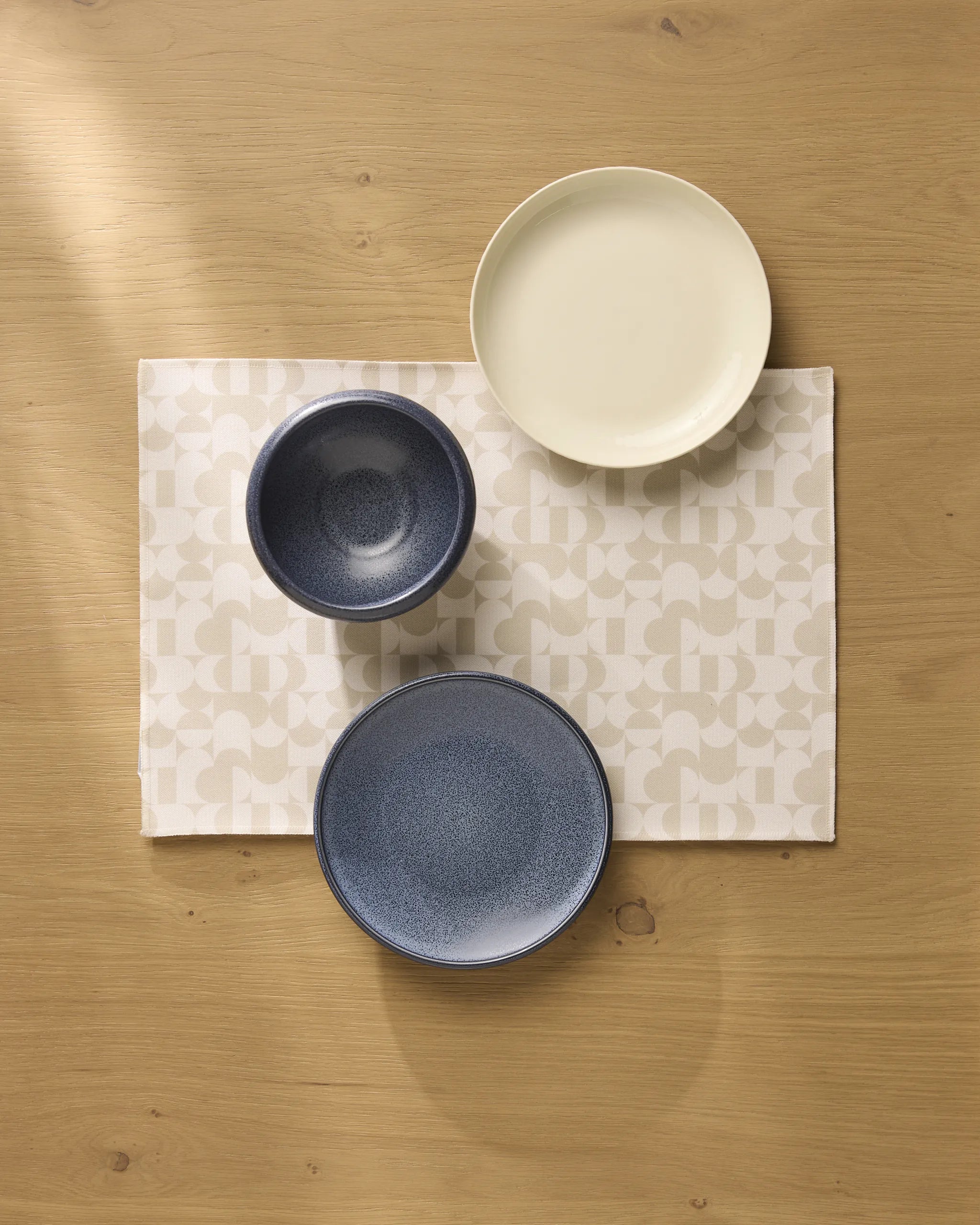 Three Circle Playground bowls by Geometry—two blue, one cream—are arranged on a patterned beige recycled placemat atop a light wooden table, viewed from above.