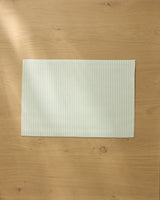 A pale green and white striped Seagrass placemat by Geometry rests on a light wooden table, with soft natural light casting gentle shadows across the surface.