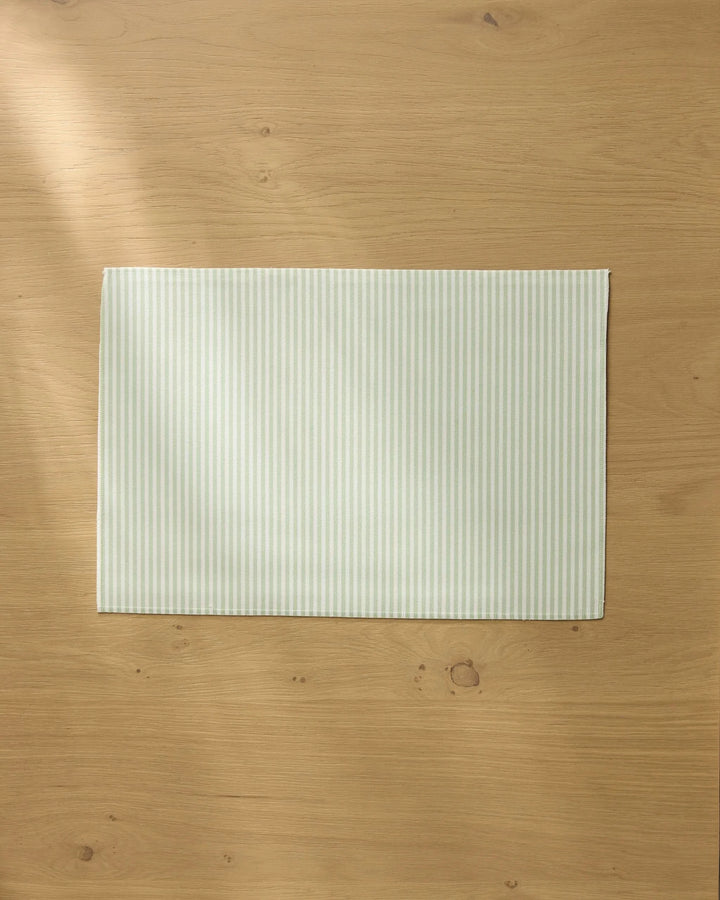 A pale green and white striped Seagrass placemat by Geometry rests on a light wooden table, with soft natural light casting gentle shadows across the surface.