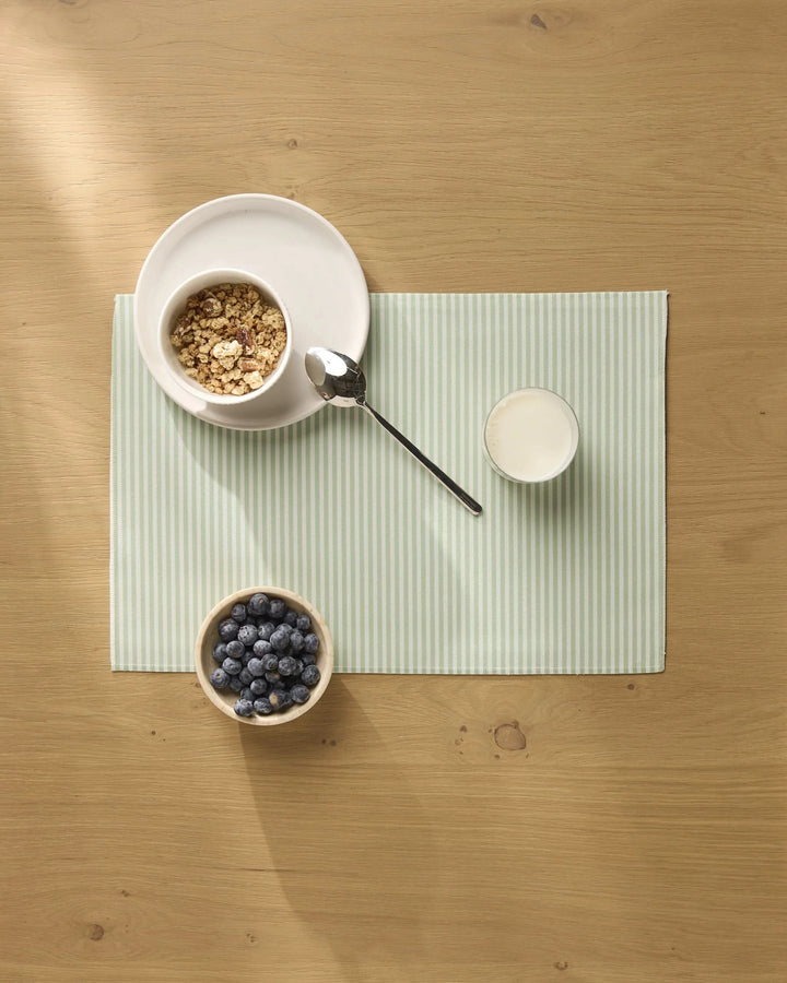 A bowl of granola, a glass of milk, and blueberries are arranged on Geometry brand Seagrass placemats with light green stripes on a wooden table.
