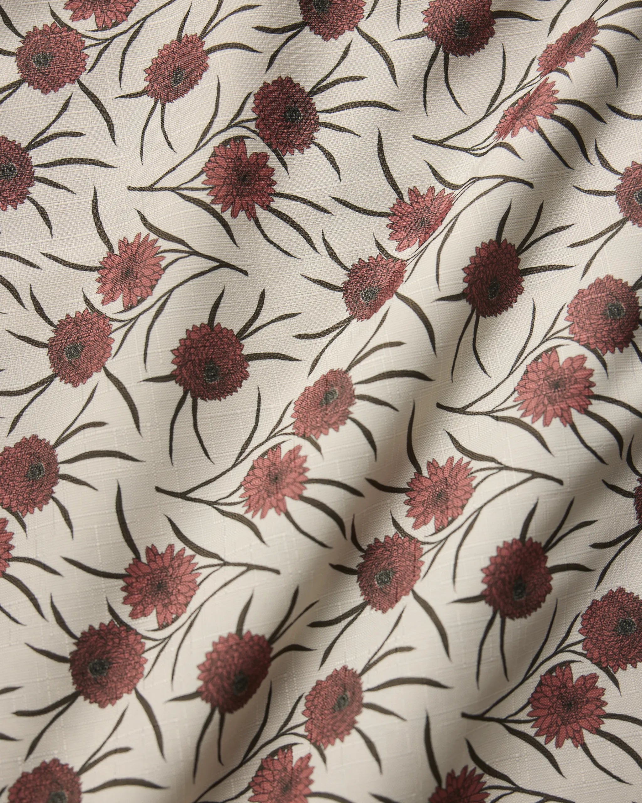 The Wild Dahlia tablecloth by Geometry is cream-colored and stain-resistant, featuring a repeating pattern of red flowers with green stems, accented by subtle folds and soft shadows.