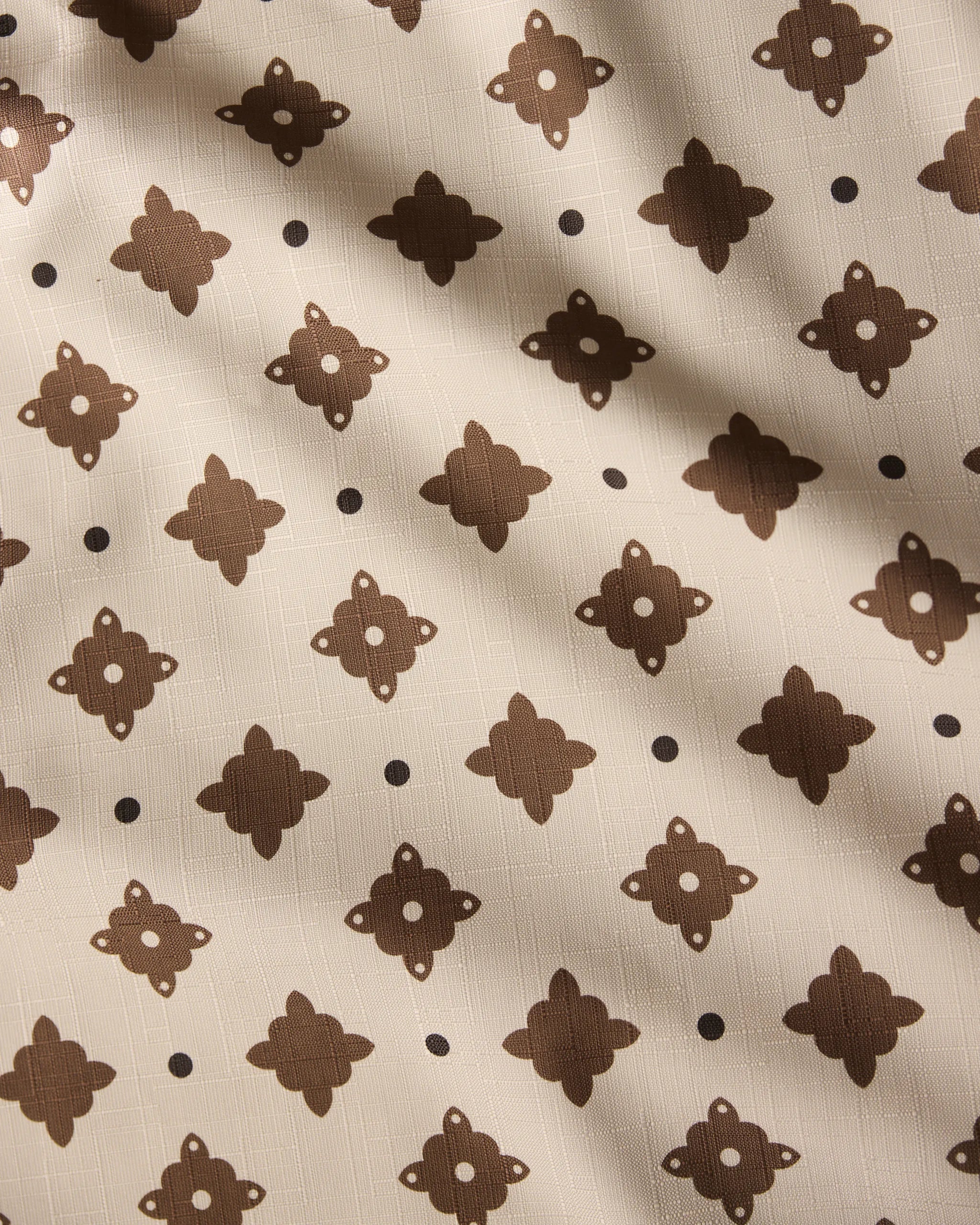 The Heritage Stamp tablecloth by Geometry features beige fabric with a repeating dark brown geometric pattern and dots, accented by subtle diagonal shadows—an eco-friendly choice perfect for modern homes.