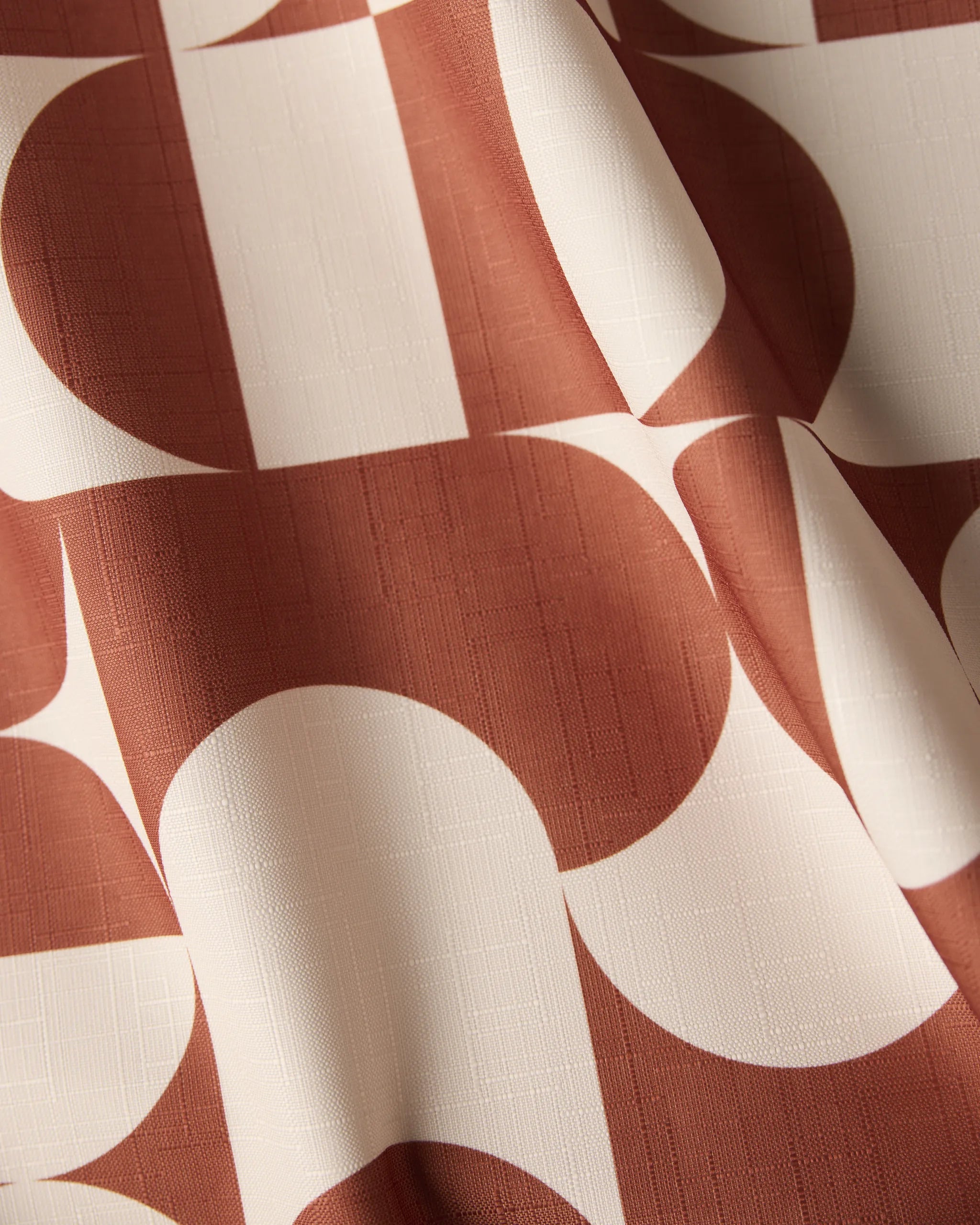 Close-up of Geometrys Circle Playground tablecloth, showcasing a sustainable fabric with a rust and off-white geometric pattern of overlapping circles and rectangles. Soft folds and shadows highlight its textured drape.
