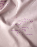A close-up of Geometrys Bubble Ditzy Dusty Rose tablecloth shows stain-resistant light pink fabric with a subtle grid, soft texture, and gentle folds, decorated with clusters of small purple dots and wavy lines.