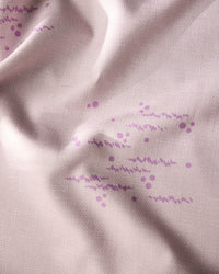 A close-up of Geometrys Bubble Ditzy Dusty Rose tablecloth shows stain-resistant light pink fabric with a subtle grid, soft texture, and gentle folds, decorated with clusters of small purple dots and wavy lines.