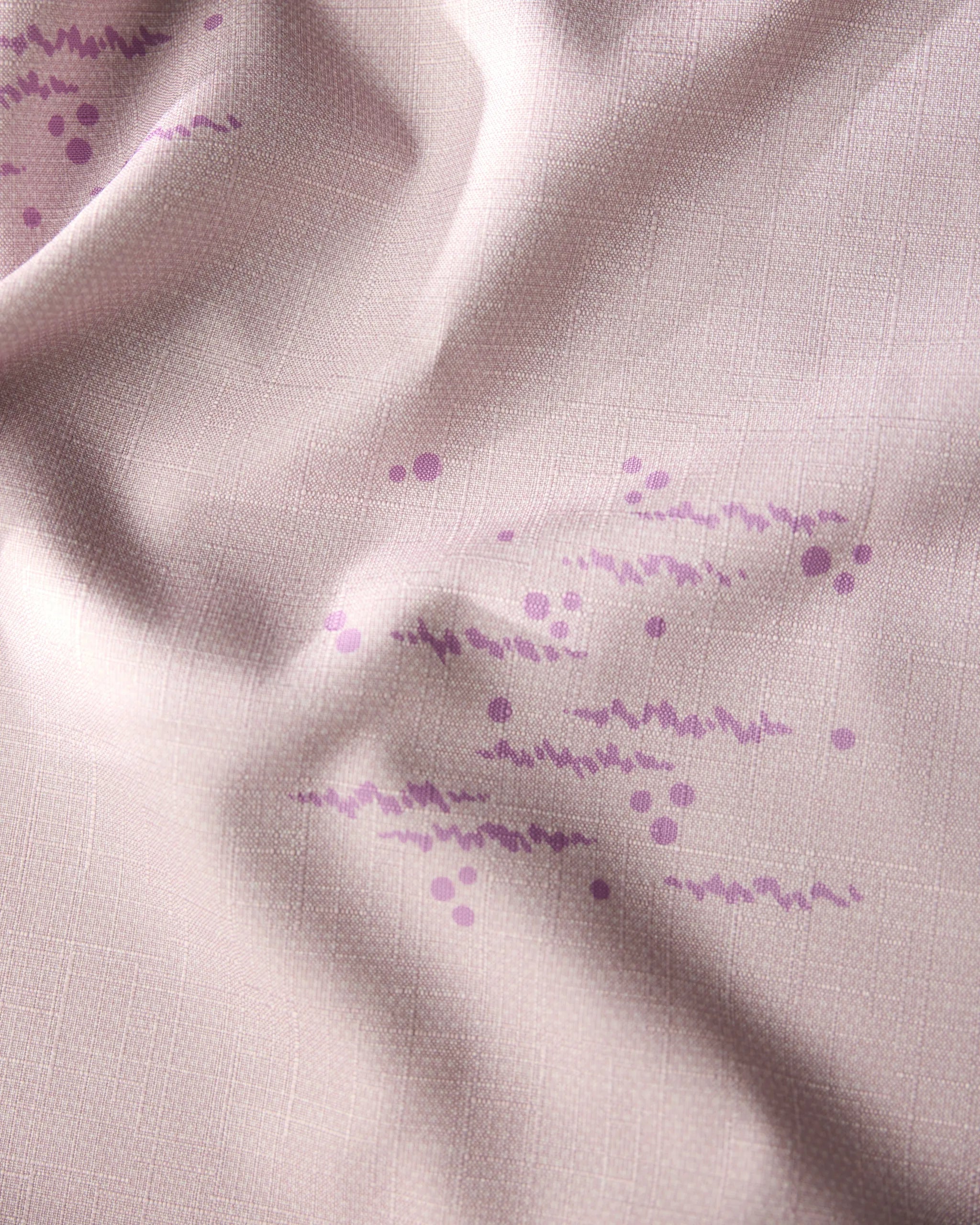 A close-up of Geometrys Bubble Ditzy Dusty Rose tablecloth shows stain-resistant light pink fabric with a subtle grid, soft texture, and gentle folds, decorated with clusters of small purple dots and wavy lines.