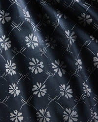 Bonnie Indigo by Geometry is a dark blue, stain-resistant tablecloth with a repeating white floral pattern of stylized flowers and intersecting diagonal lines. Its slightly textured fabric drapes elegantly, creating soft folds and shadows.