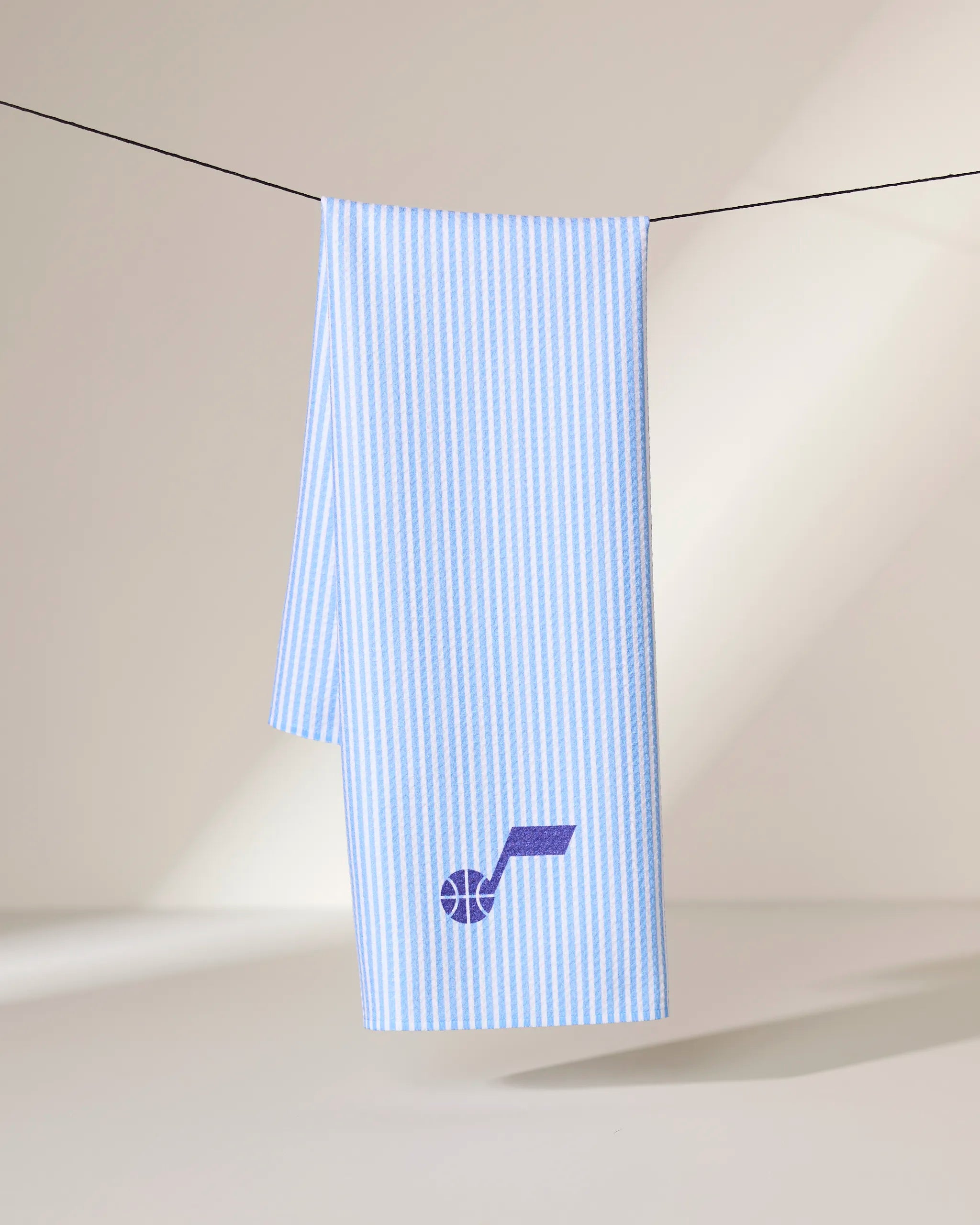 The Geometry Jazz Classic towel, featuring light blue and white vertical stripes with a purple basketball and note logo, hangs over a black clothesline against a neutral background.