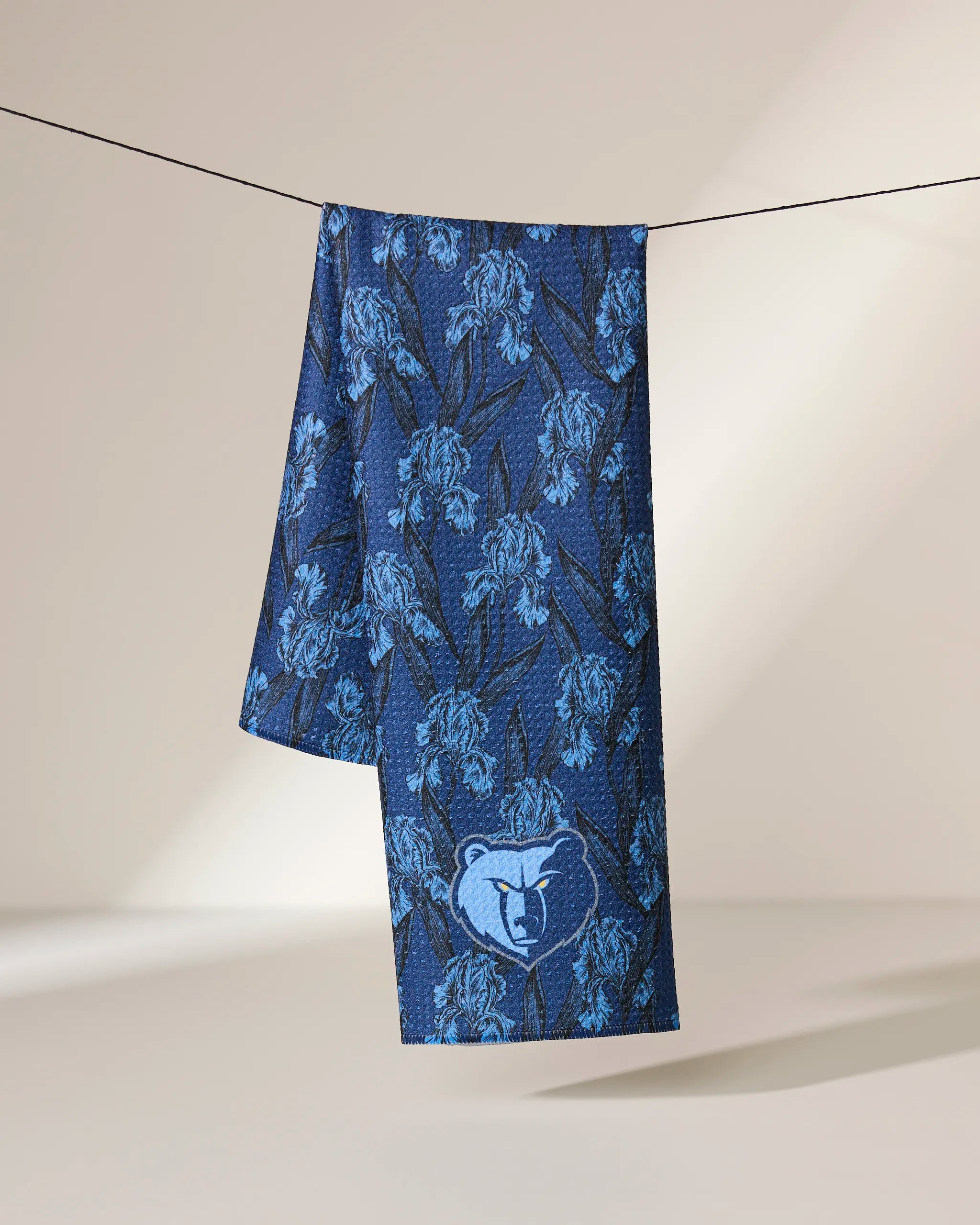 The Grizzlies Floral towel by Geometry, featuring a blue floral pattern and bear logo, hangs over a clothesline against a neutral background with soft lighting.