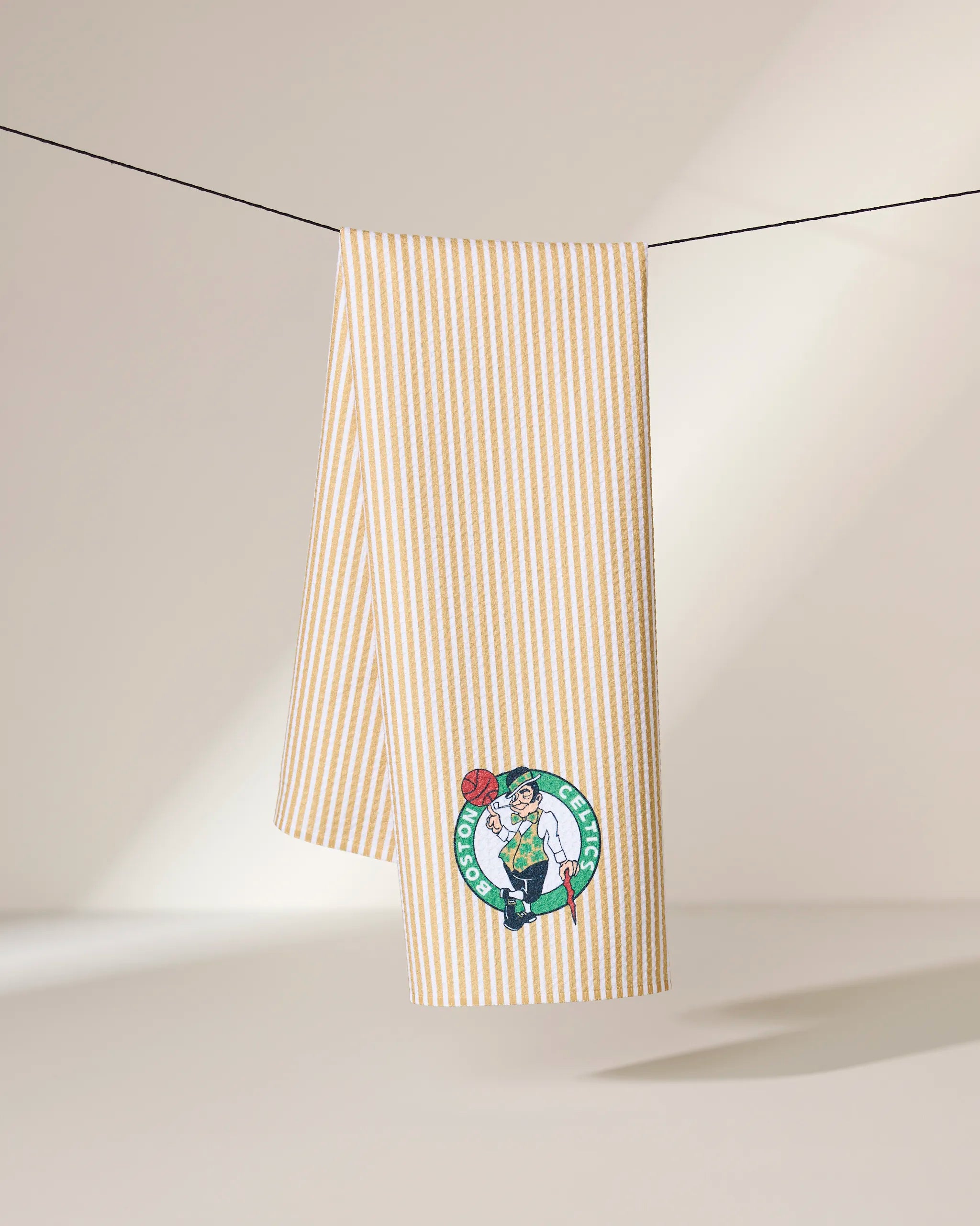 A yellow and white striped Celtics Classic towel by Geometry hangs on a line, featuring the Boston Celtics logo with the team’s mascot in the bottom corner. The neutral background is softly lit.