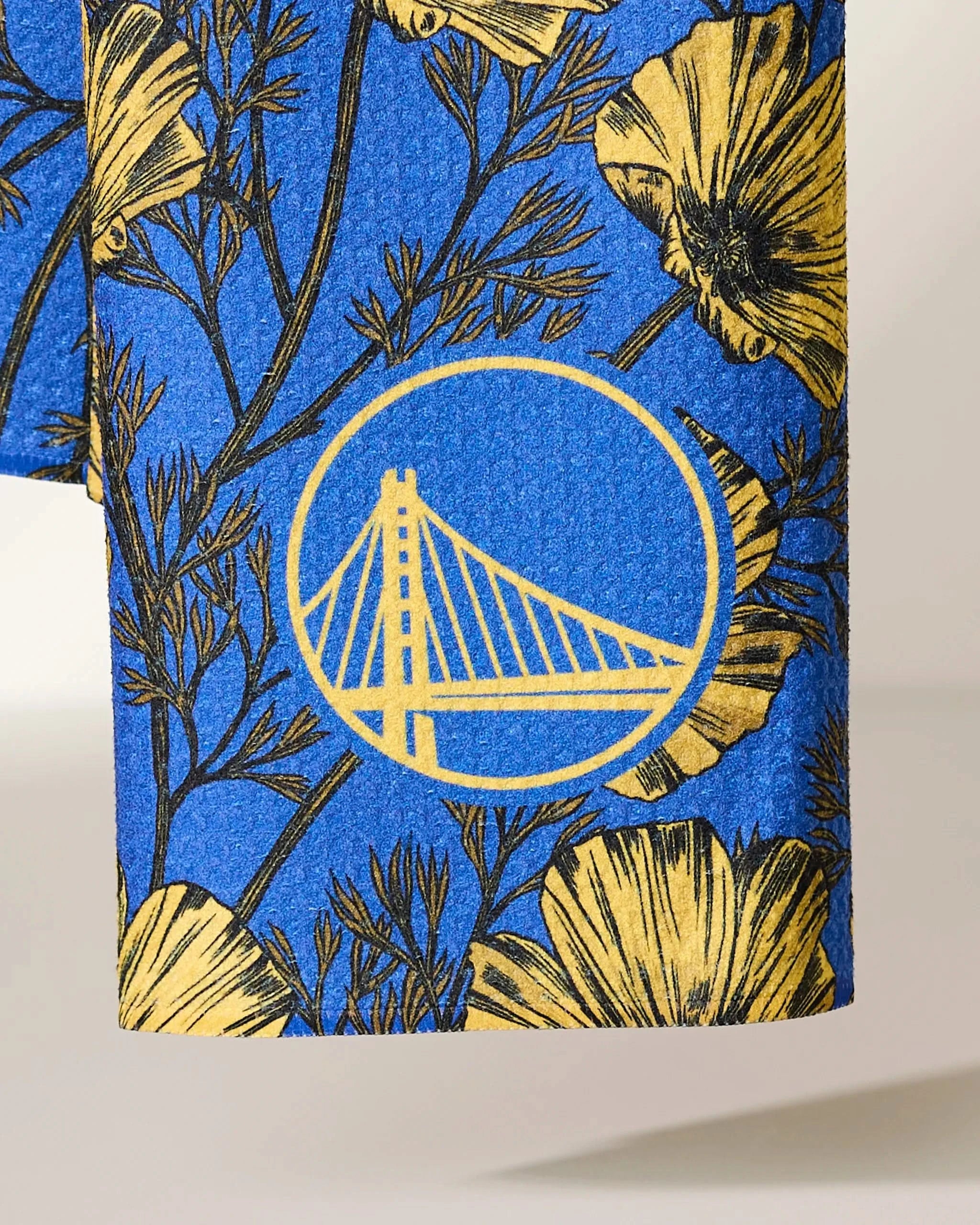 A close-up of blue Warriors Floral fabric by Geometry, featuring yellow and black floral patterns and a yellow suspension bridge outline in a circle, resembling the Golden State Warriors logo.