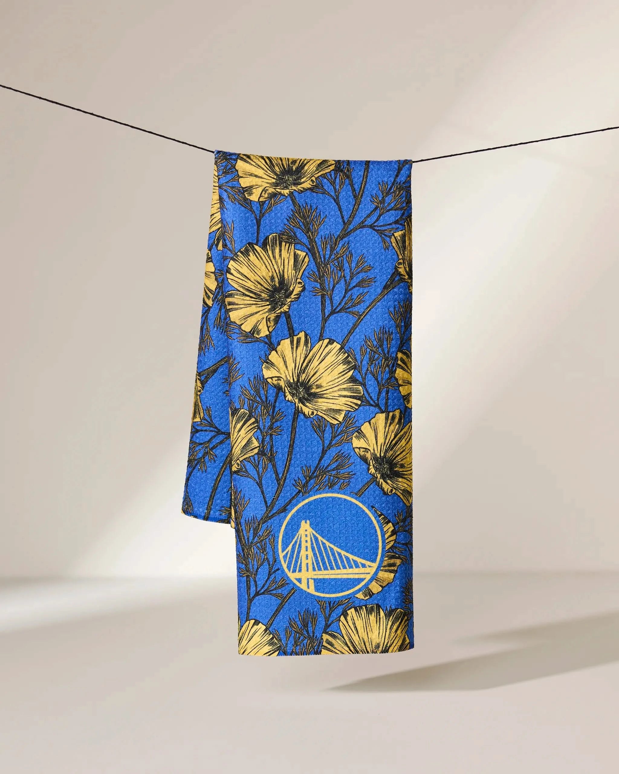 The Geometry Warriors Floral towel, featuring a yellow floral pattern and Golden State Warriors bridge logo on blue, hangs on a black line against a light background.