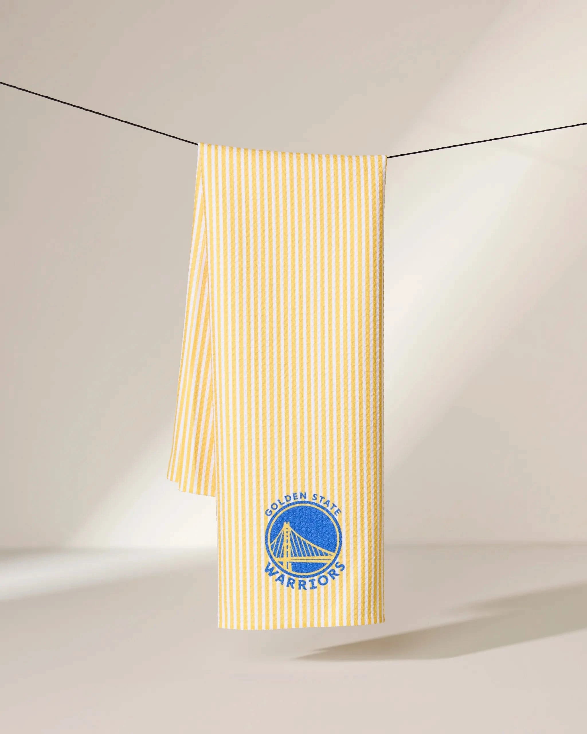 The Geometry Warriors Classic towel, featuring yellow and white stripes and the Golden State Warriors logo, hangs on a black string against a neutral background.