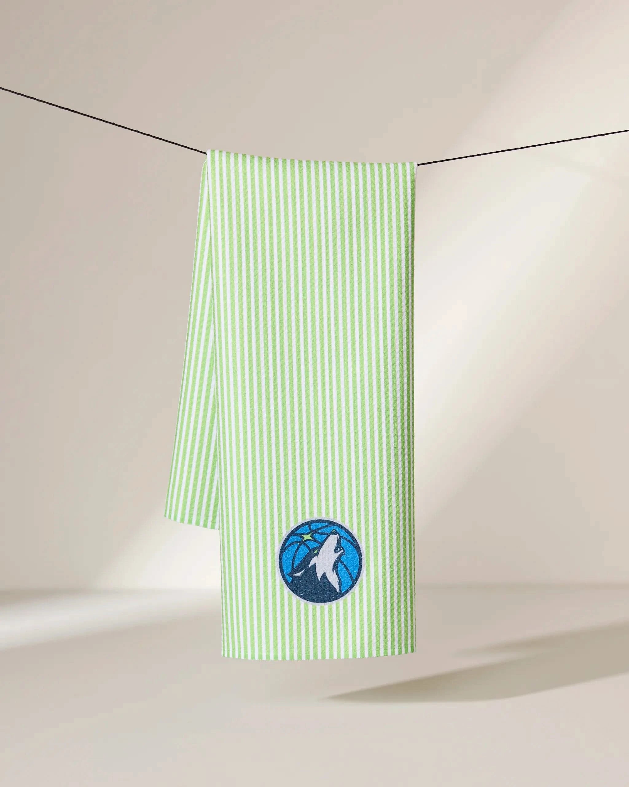 The Geometry Timberwolves Classic towel, green and white striped, hangs on a black line. It showcases an embroidered blue and white basketball logo with a wolf’s head against a softly lit, minimalistic background.