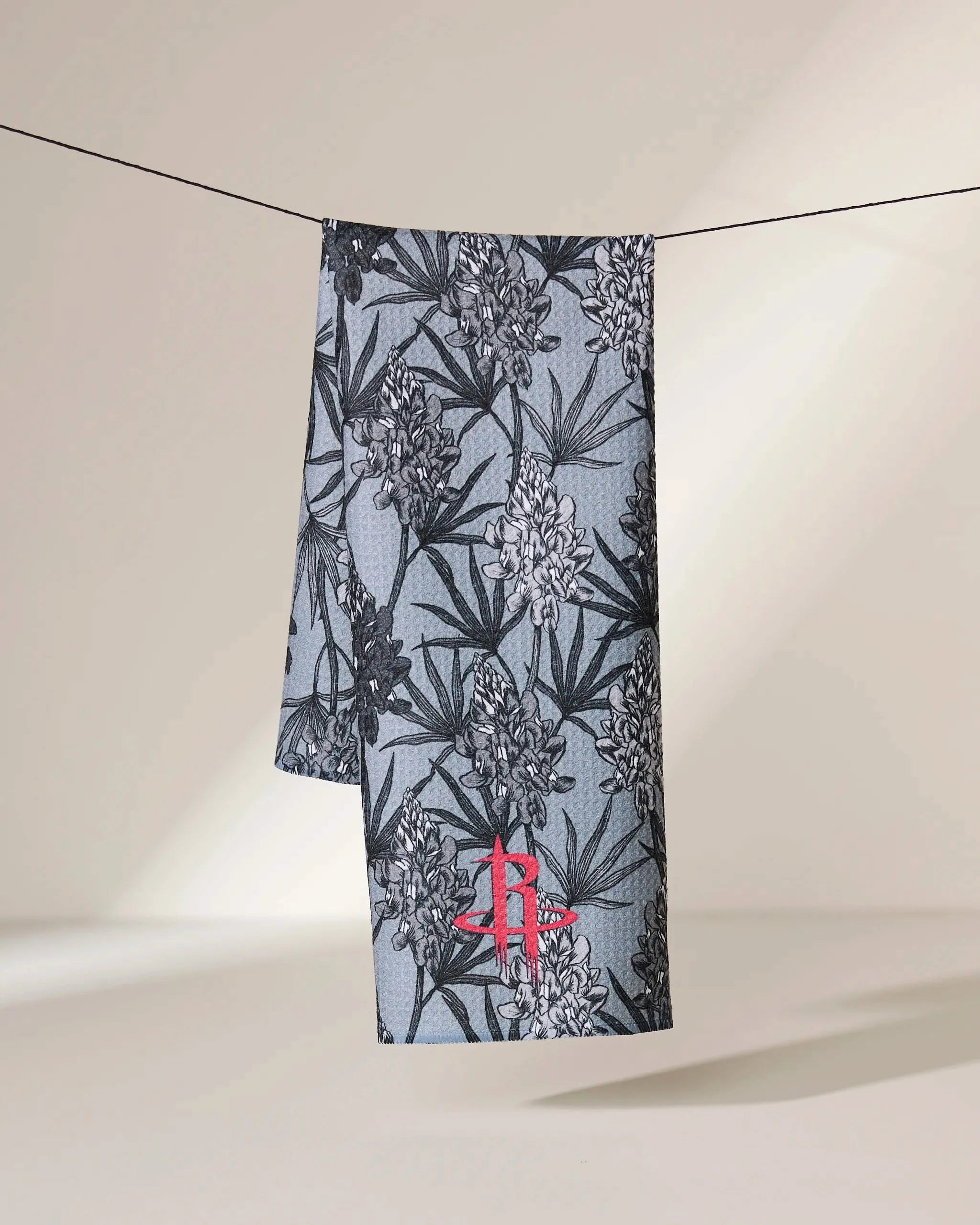 The Geometry Rockets Floral towel, featuring a black floral pattern on gray fabric and the red Houston Rockets logo near the bottom, hangs on a line against a neutral background with soft lighting.
