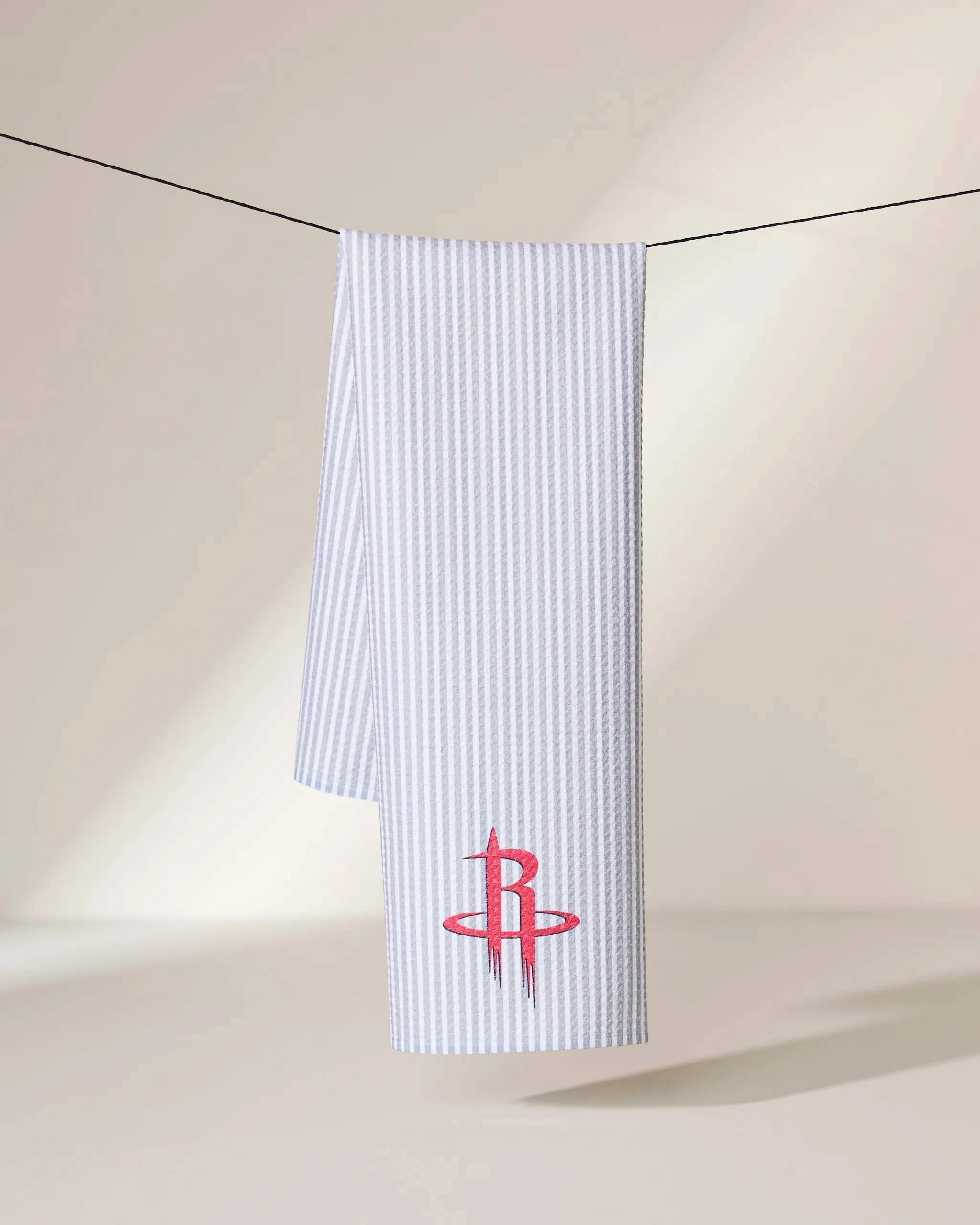 The Geometry Rockets Classic towel, featuring a red Houston Rockets logo on white ribbed fabric, hangs on a black line against a neutral beige background.