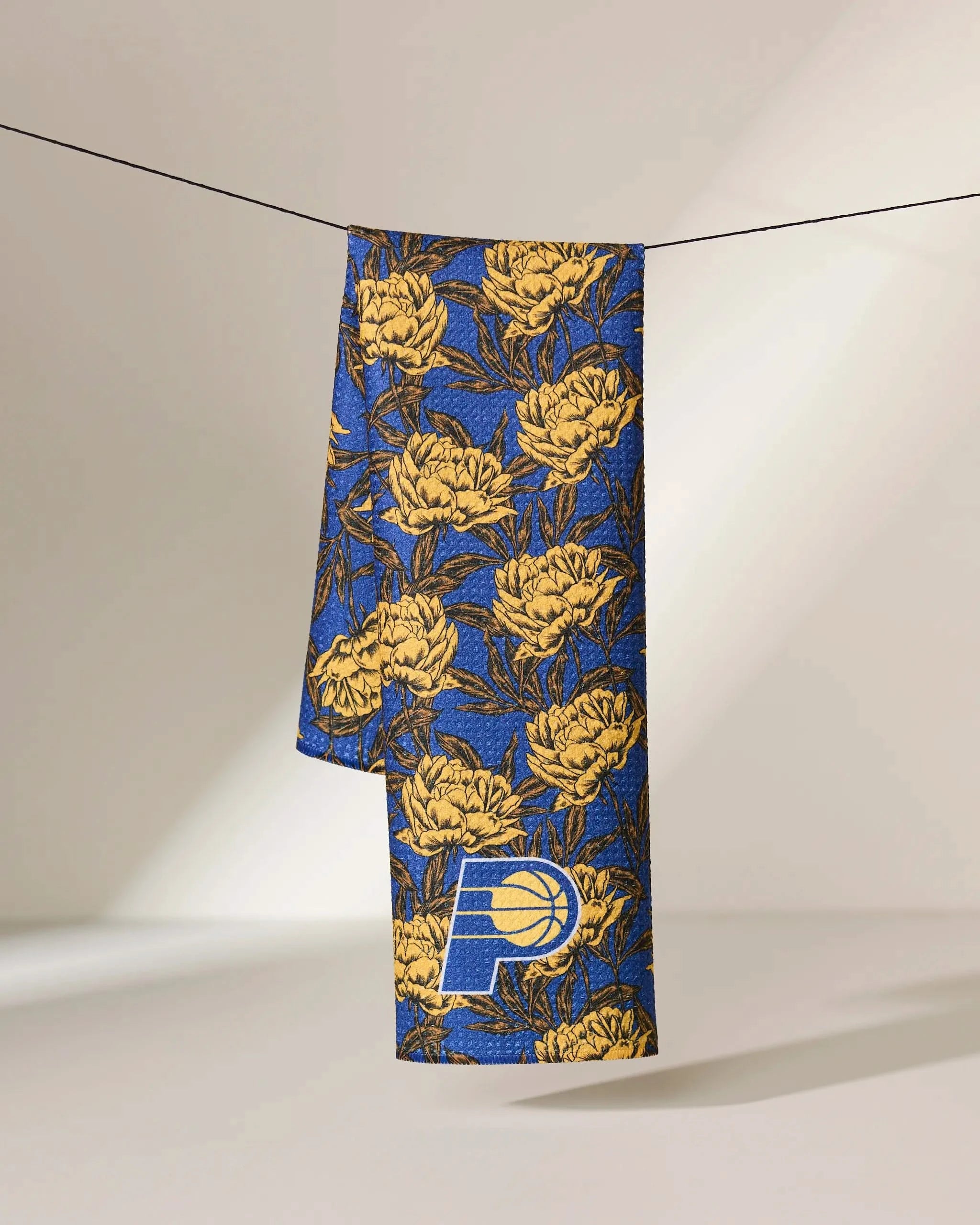 The Geometry Pacers Floral towel, featuring blue and gold floral patterns with the Indiana Pacers logo, hangs on a line against a softly lit background.