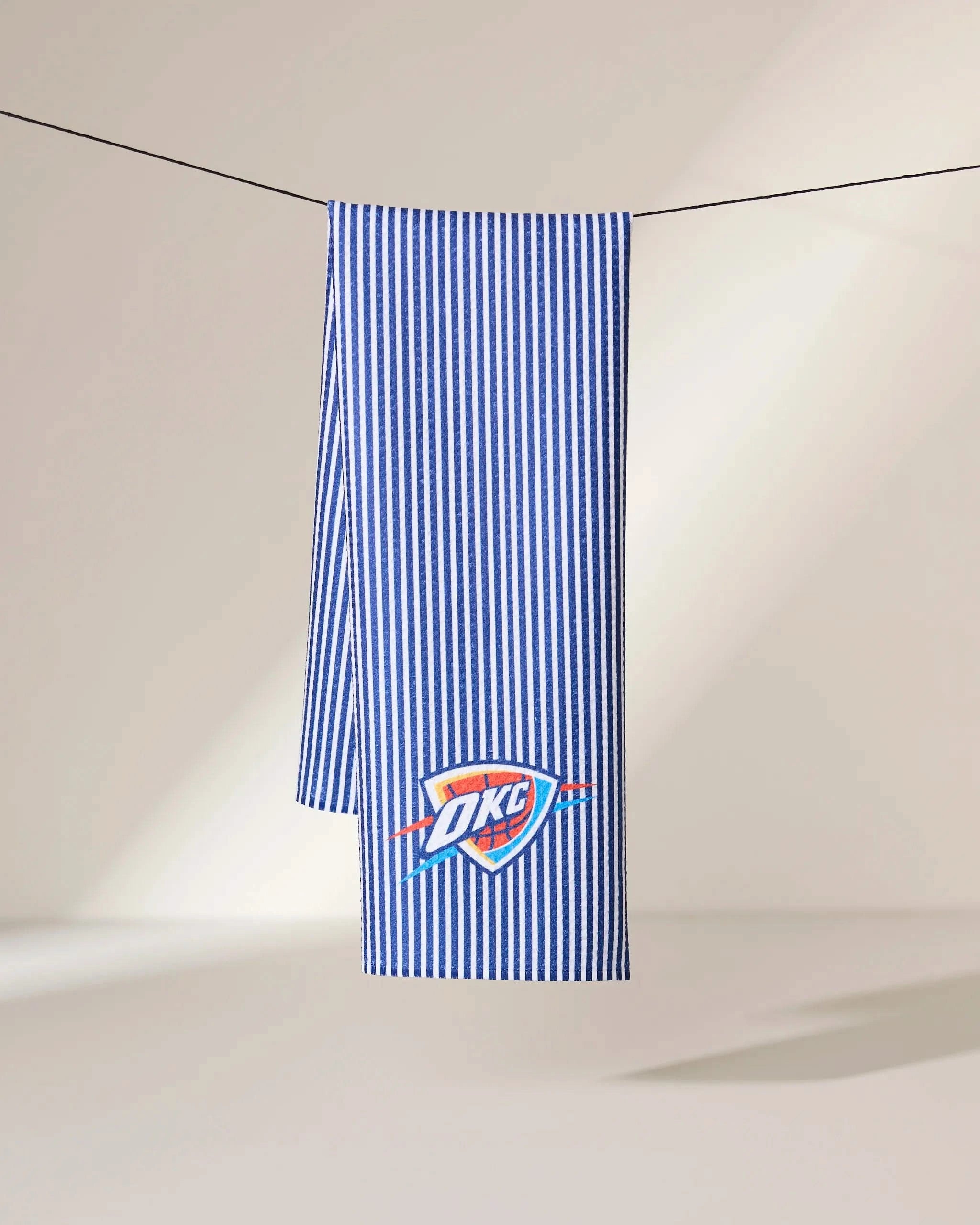 The Geometry OKC Classic towel features blue and white vertical stripes with the Oklahoma City Thunder logo, shown hanging on a black clothesline against a neutral background.