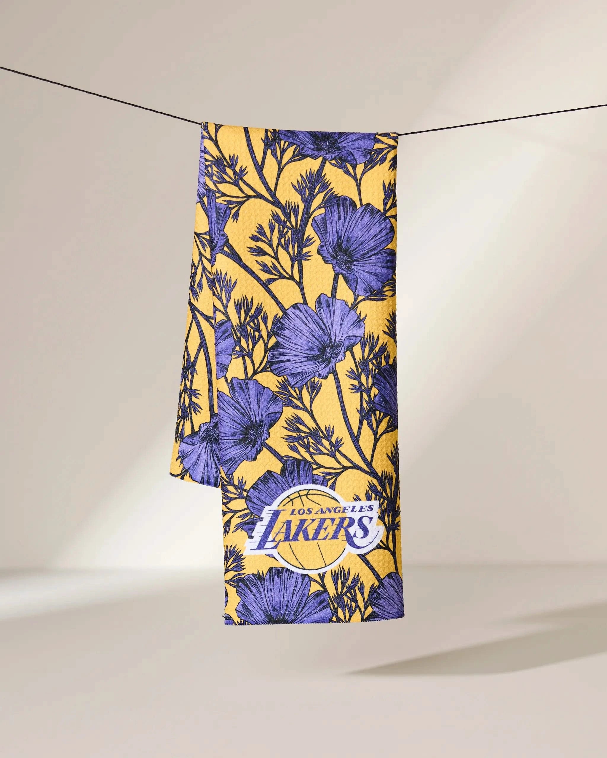 The Geometry Lakers Floral towel, featuring purple floral designs and the Los Angeles Lakers logo, hangs over a slim black line against a plain, light background.