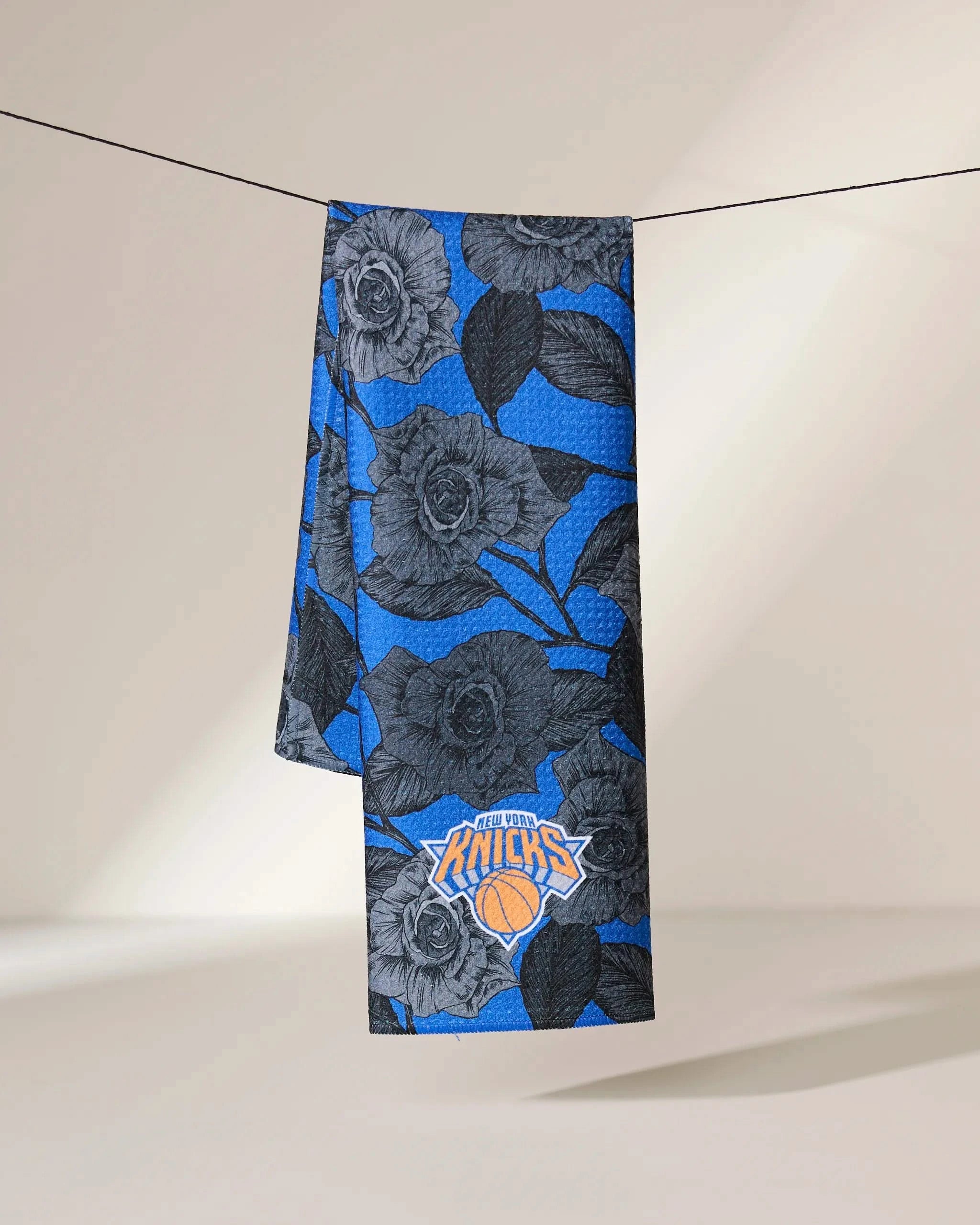 The Geometry Knicks Floral towel, featuring a black floral pattern and New York Knicks logo, hangs draped over a black line against a neutral background.