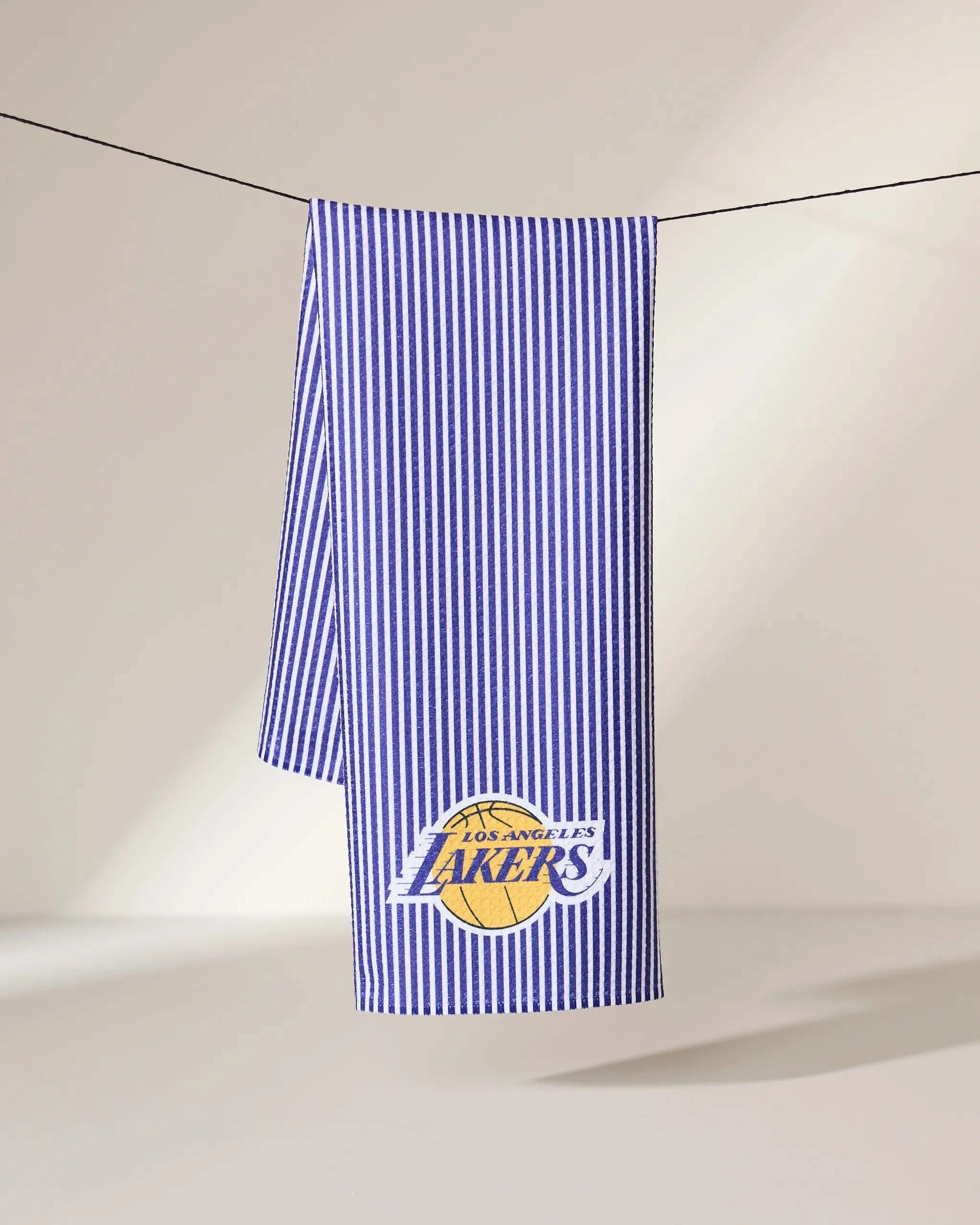 The Geometry Lakers Classic towel, in blue and white stripes, hangs on a line with the Los Angeles Lakers logo in yellow and purple displayed in the bottom corner.