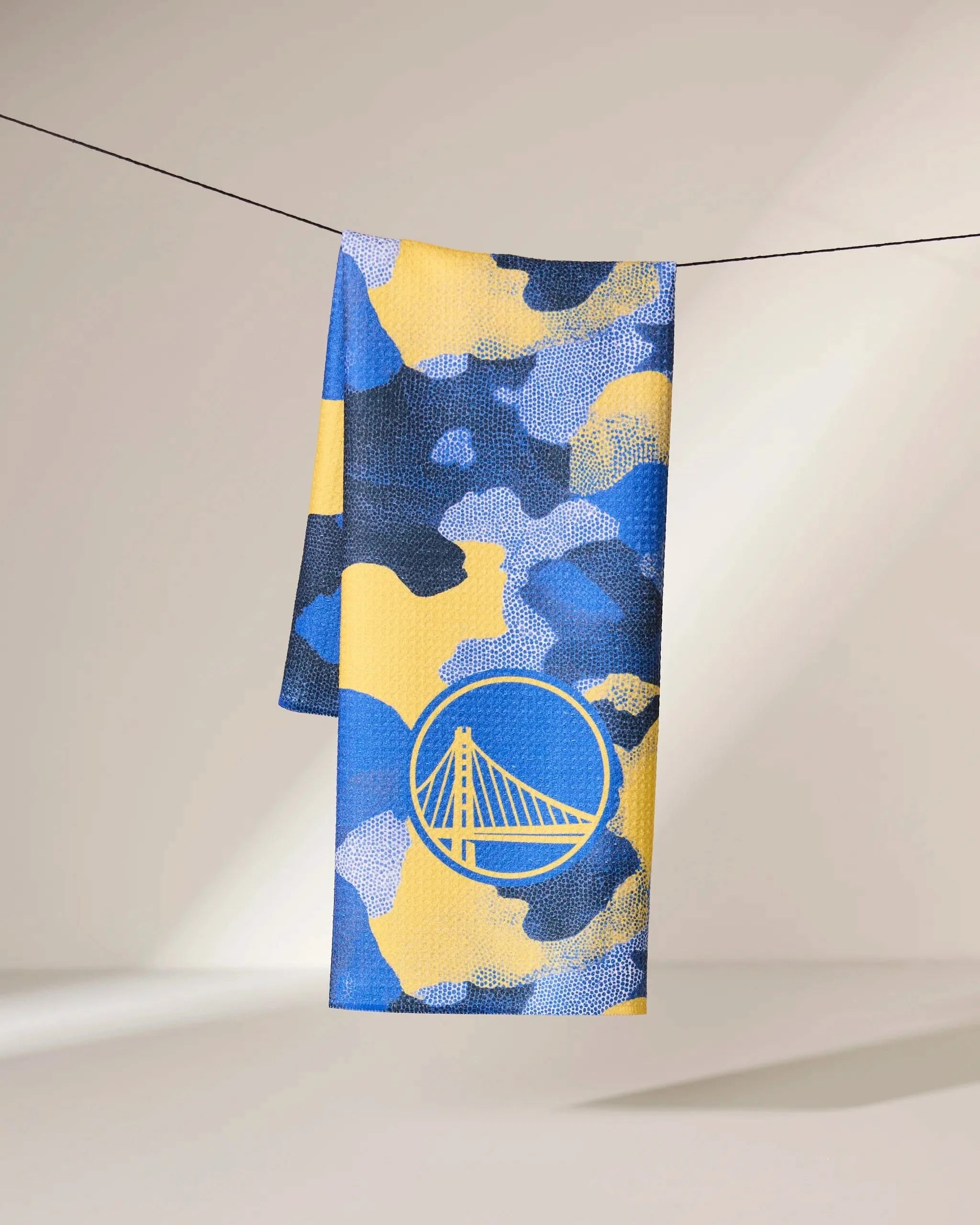 The Geometry Warriors Camo towel, featuring a blue and yellow pattern, hangs neatly over a black clothesline against a neutral background.