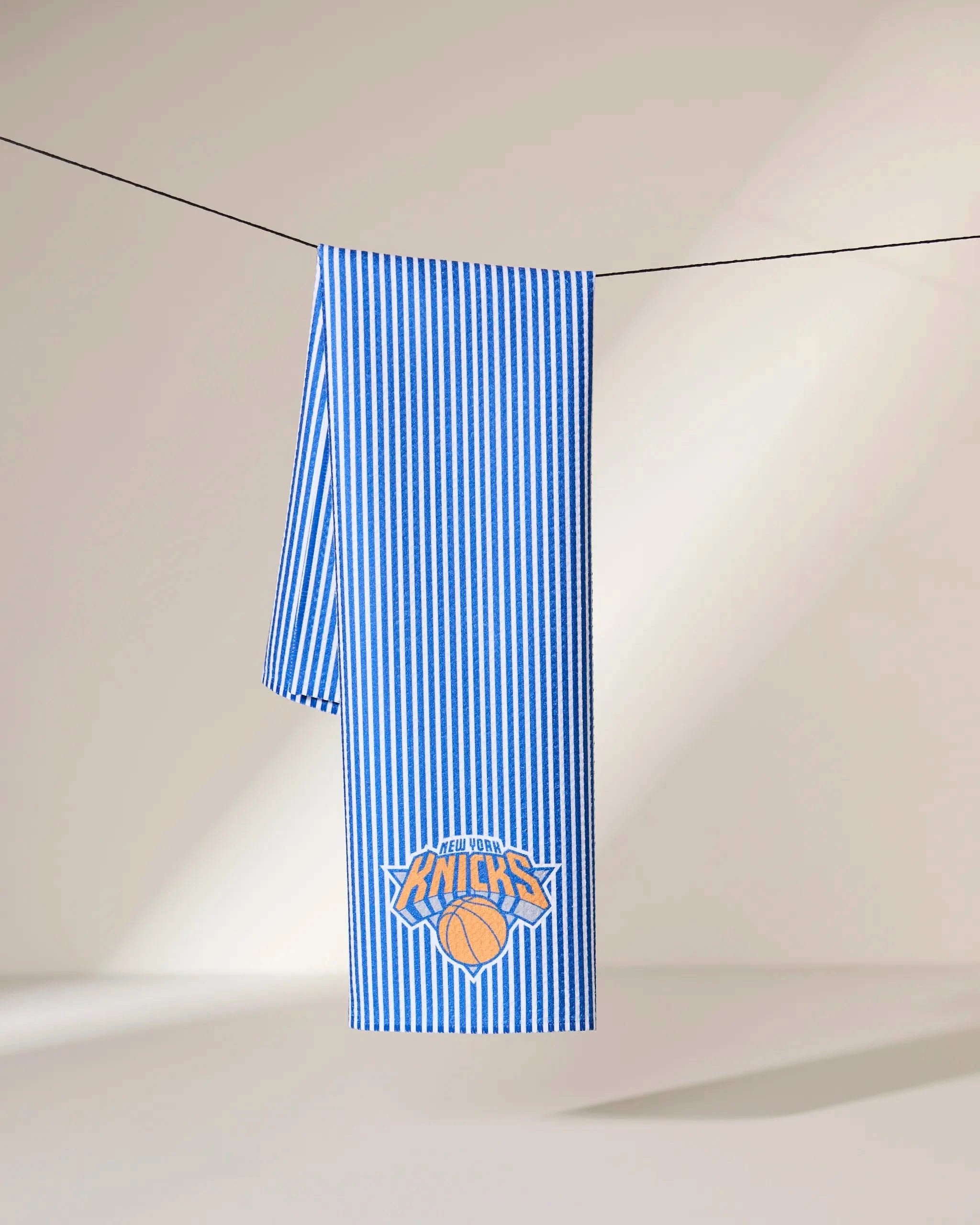 The Geometry Knicks Classic towel, featuring blue and white vertical stripes and the New York Knicks logo, is draped over a black line against a neutral background.