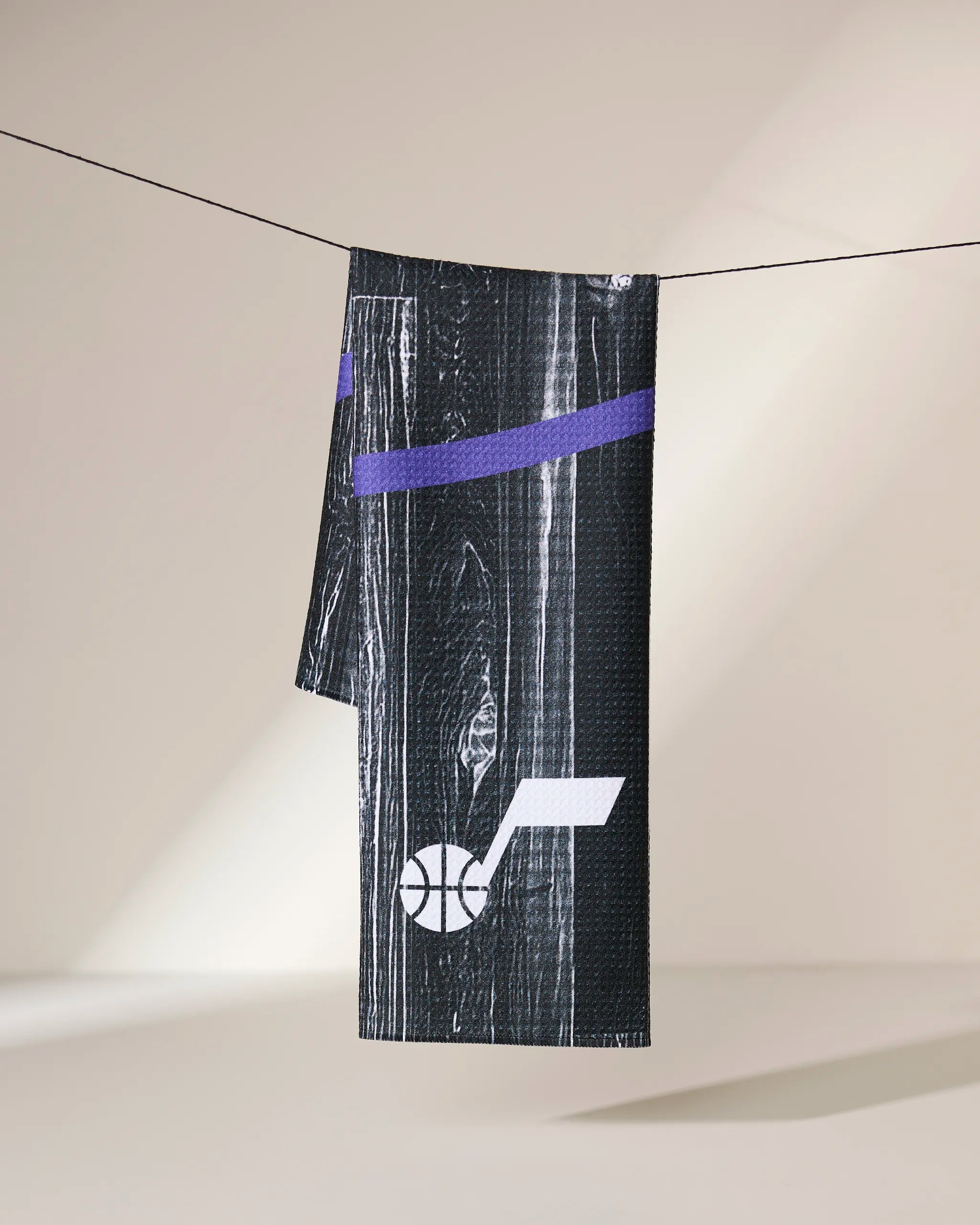 A Geometry Jazz Zone black sports towel with a purple stripe and white Utah Jazz basketball logo hangs on a clothesline against a neutral background.