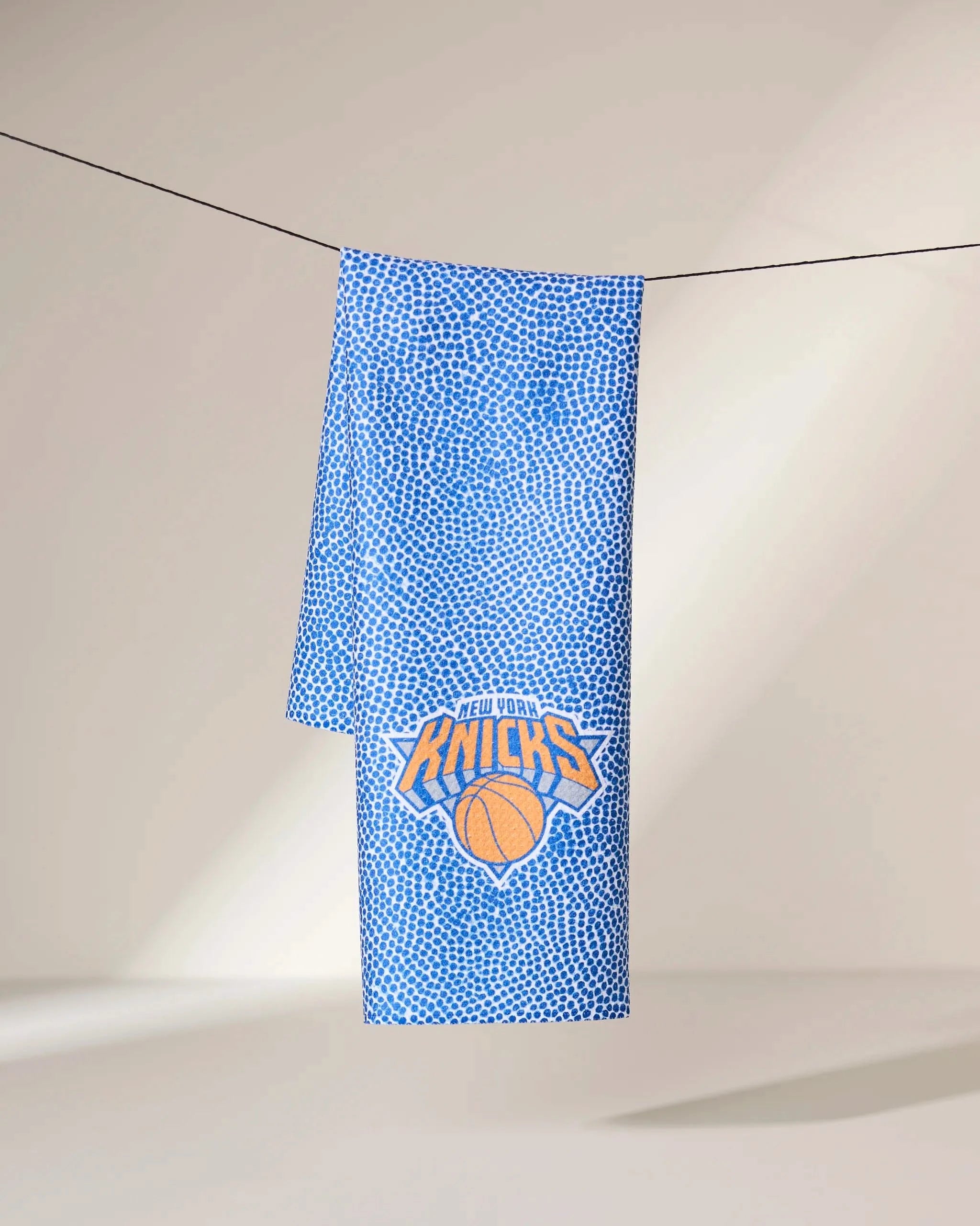 A blue dotted Geometry Knicks Grip towel hangs on a clothesline, featuring the New York Knicks basketball team logo in orange and blue near the bottom.