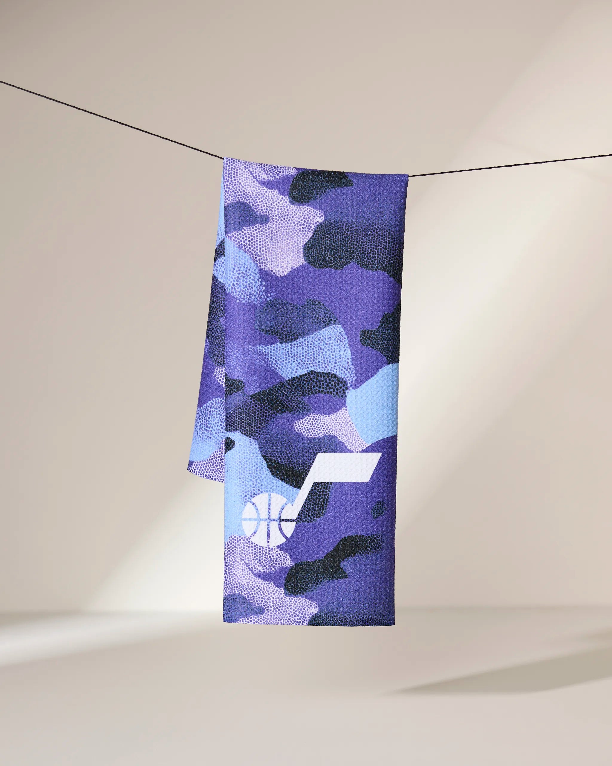 The Geometry Jazz Camo towel, featuring a purple camouflage pattern and white basketball with musical note logo, hangs on a black string against a softly lit background.