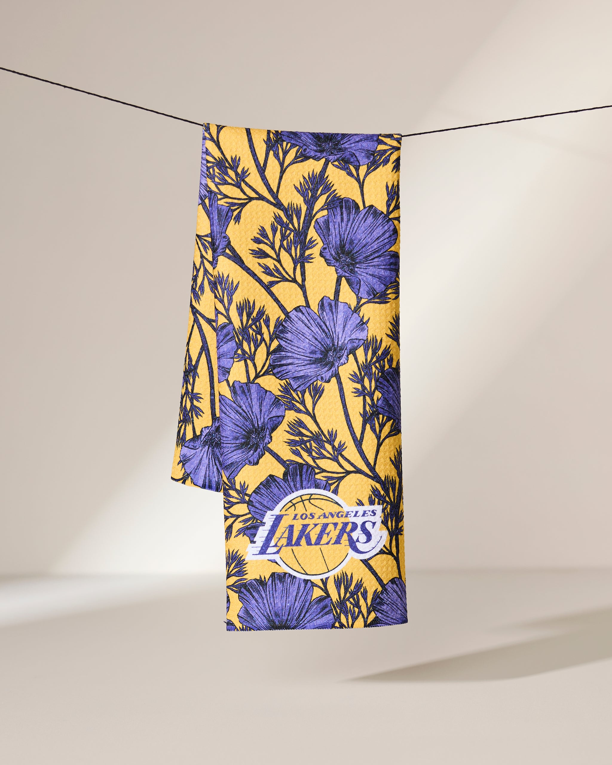 The Geometry Lakers Floral towel, featuring purple floral patterns and the Los Angeles Lakers logo in yellow, hangs neatly over a black string against a neutral background.