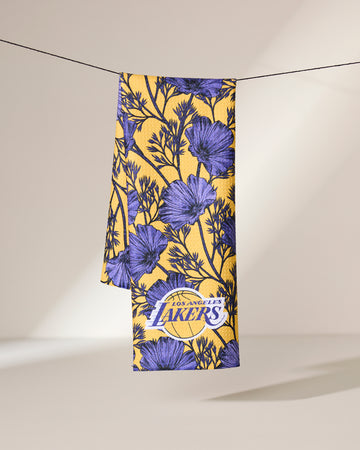 The Geometry Lakers Floral towel, featuring purple floral patterns and the Los Angeles Lakers logo in yellow, hangs neatly over a black string against a neutral background.