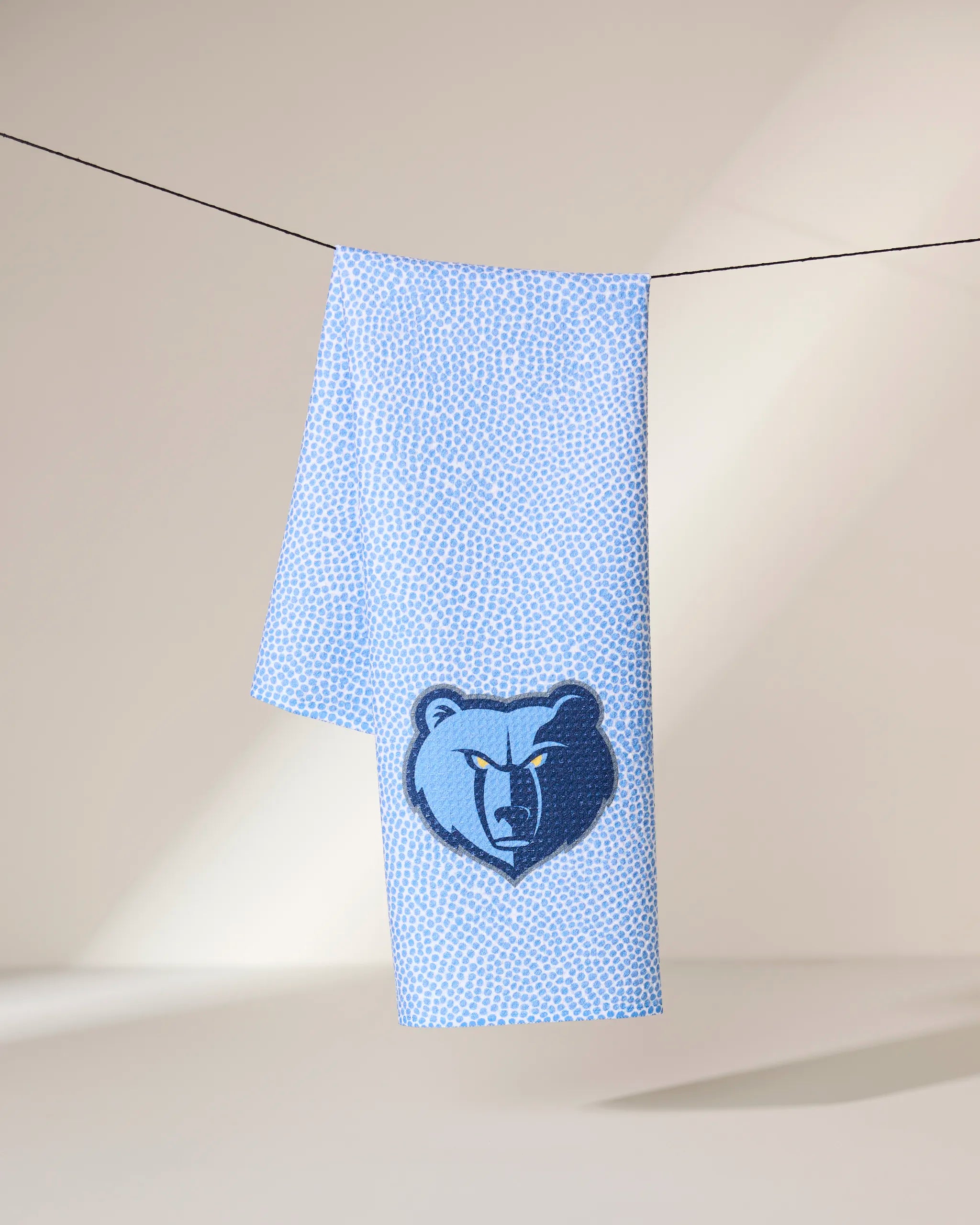 A light blue Geometry Grizzlies Grip towel with a subtle dotted pattern hangs on a line, featuring a large dark blue Memphis Grizzlies bear logo on one end against a softly lit neutral background.