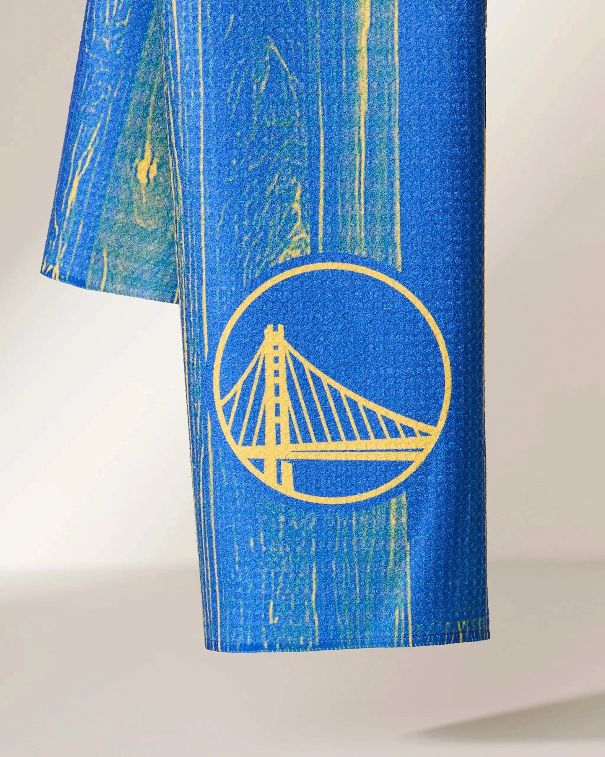 A close-up of Geometrys Warriors Zone blue fabric features a yellow bridge graphic inspired by the Warriors logo, with a subtle textured pattern and yellow streaks.